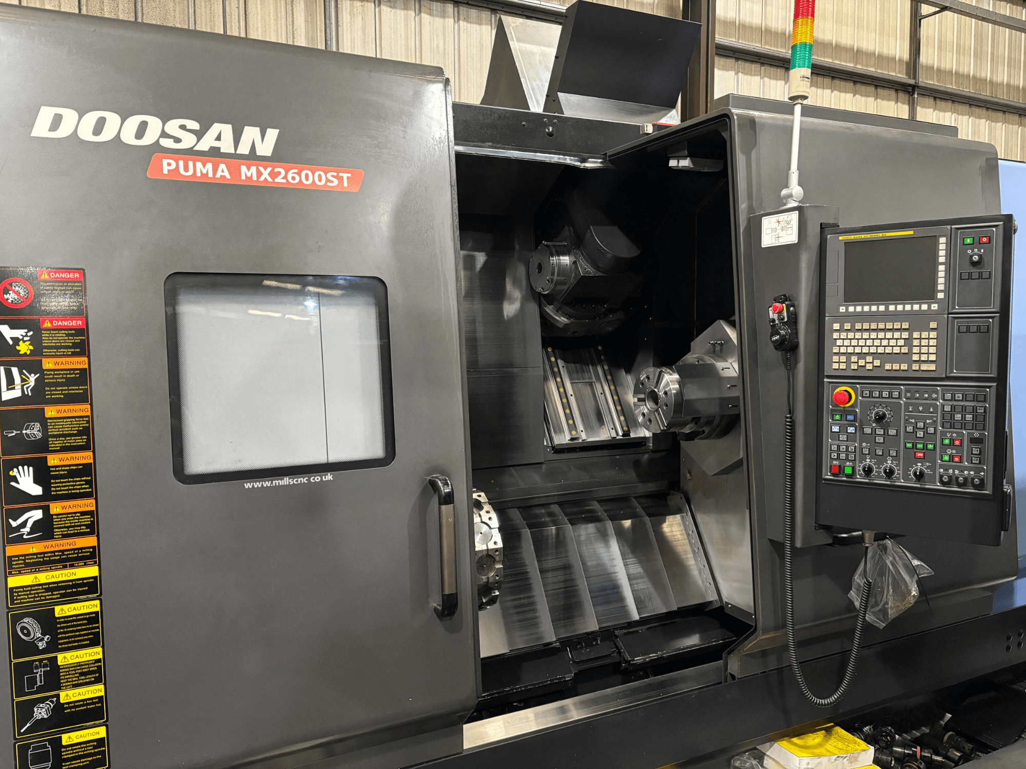 Doosan PUMA MX2600ST CNC-svarv, framifrån, visar kontrollpanelen, spindeln och bearbetningsområdet med synliga säkerhetsvarningar.