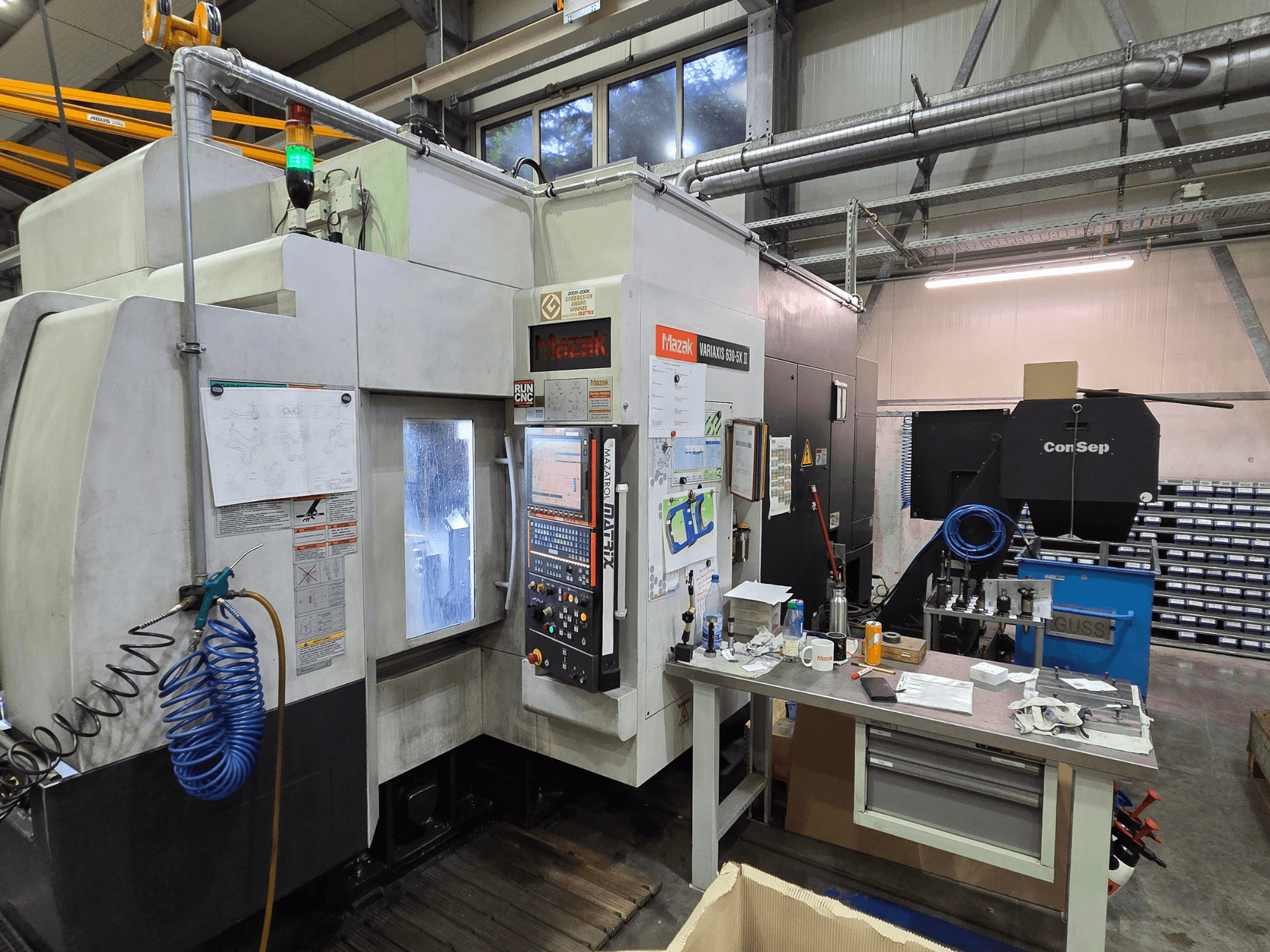 Mazak Variaxis 630-5X CNC-maskin i en verkstad, framifrån med kontrollpanel, slang och tillbehör på ett sidobord.