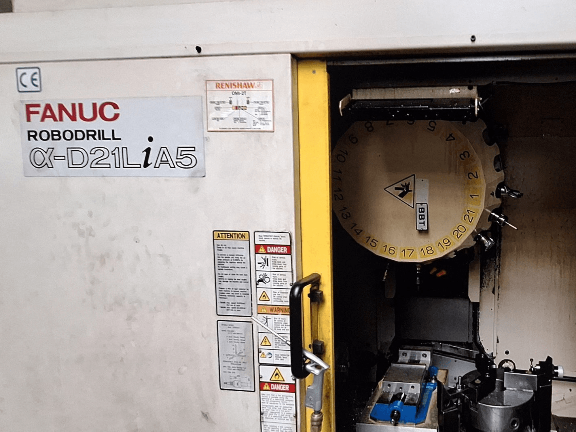 FANUC Robodrill α-D21LiA5, vy framifrån. Visar verktygskarusell och bearbetningsområde med klämmor och varningar på dörren.