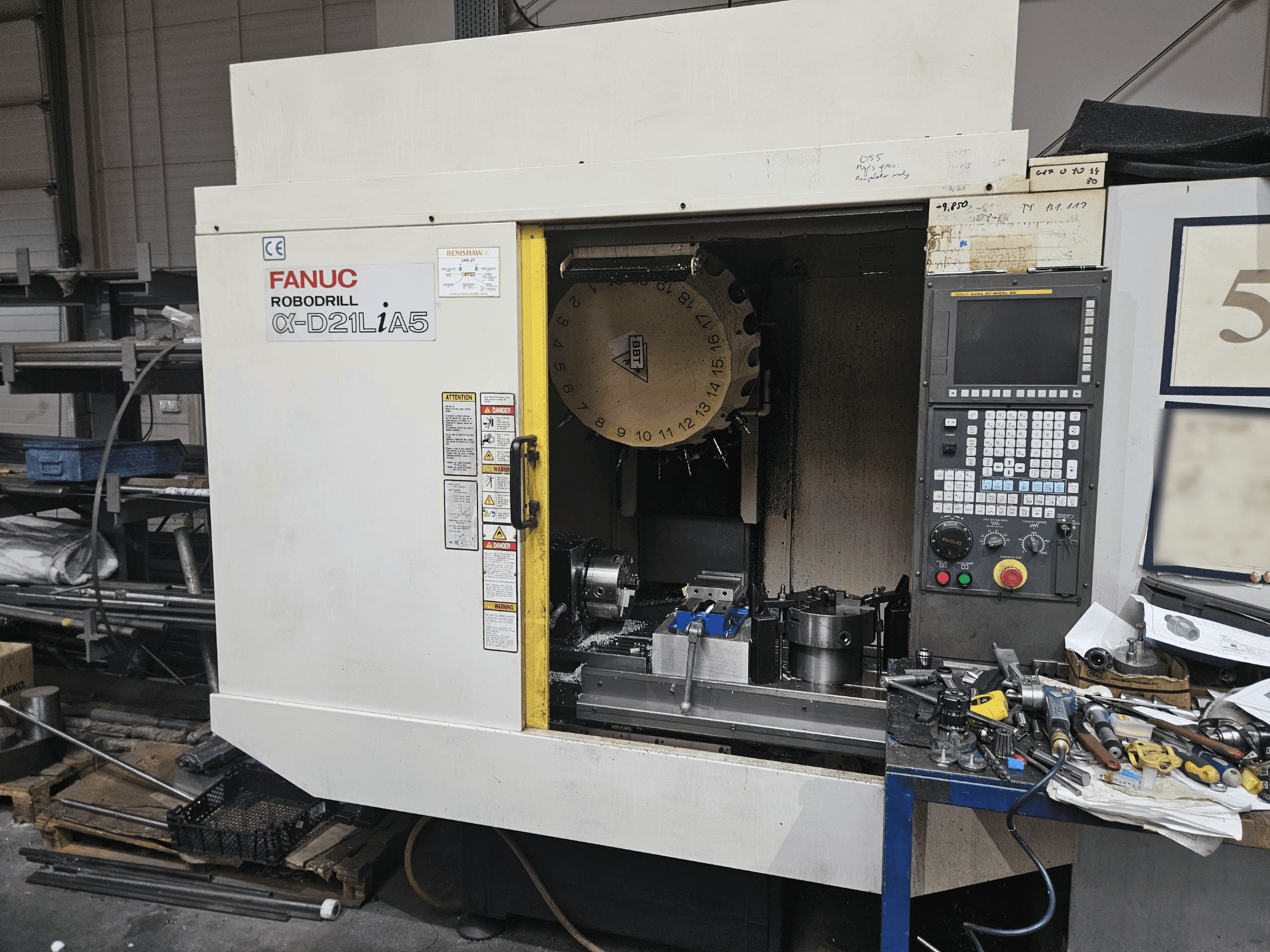 FANUC Robodrill α-D21LiA5, vy framifrån som visar verktygshållare och kontrollpanel, omgiven av verktyg och utrustning i en verkstad.