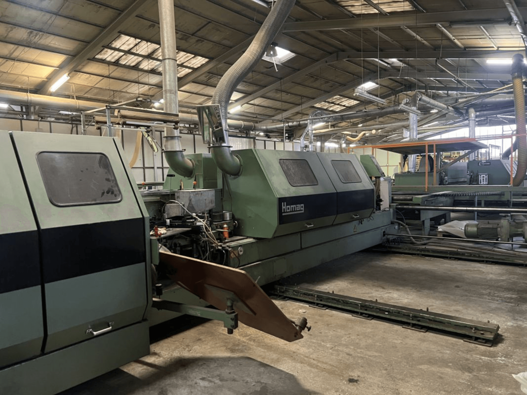 Homag industrimaskin i grönt, sidovy, med operativa komponenter och kontrollpanel synliga i en lagermiljö.