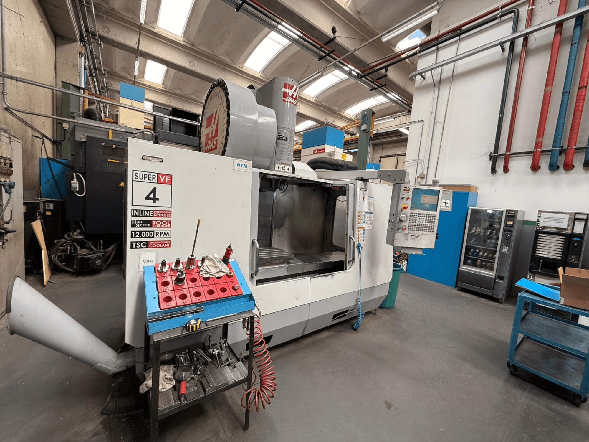 HAAS Super VF 4 CNC-fräsmaskin med kontrollpanel, spindel på 12.000 varv/min och verktyg på ett blått förvaringsställ, vy från sidan.