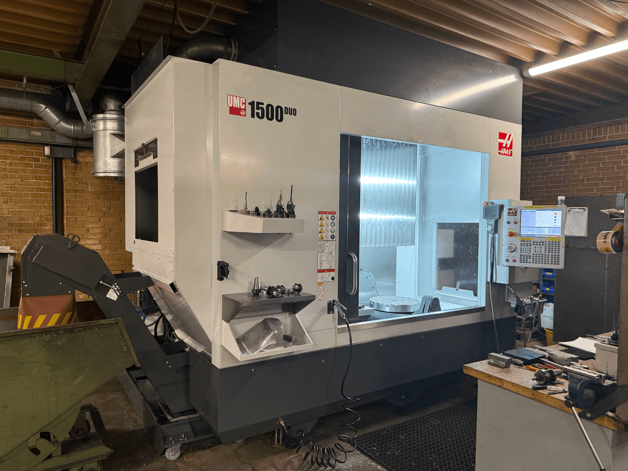 Haas UMC 1500DUO CNC-maskin, framifrån, har ett stort fönster, kontrollpanel och verktygshållare på en hylla.