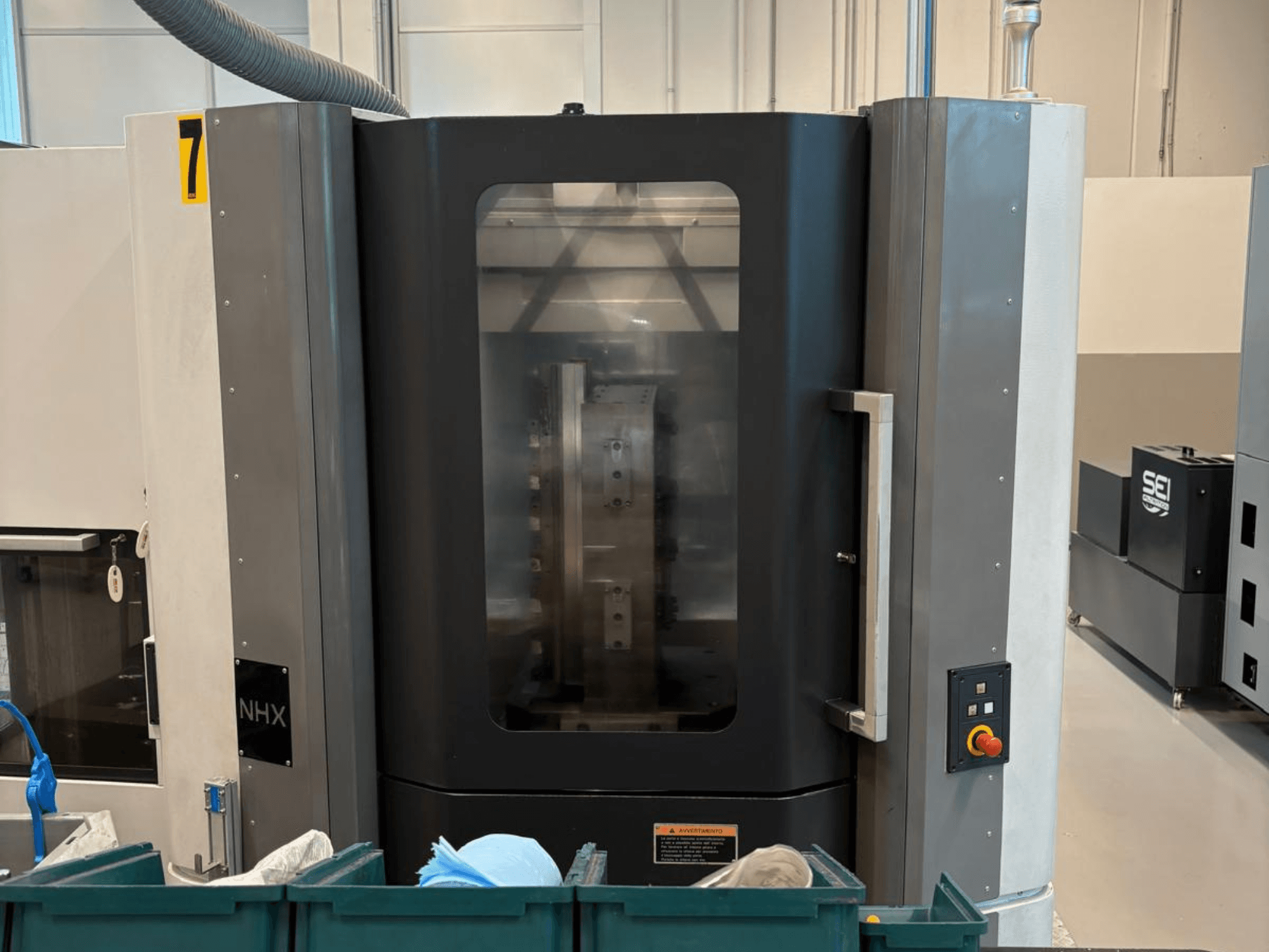 NHX CNC-maskin framifrån, med kontrollpanel och tydlig display; placerad i en verkstad med gröna soptunnor i närheten.