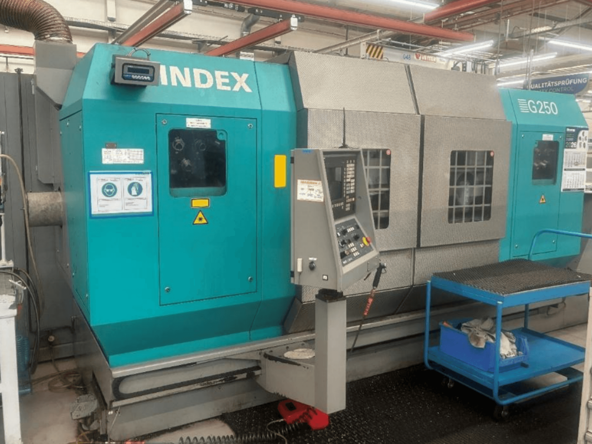 Horisontell svarv INDEX G250, framifrån sett med kontrollpanelen och metallhöljet, försedd med säkerhetsetiketter och en blå vagn.