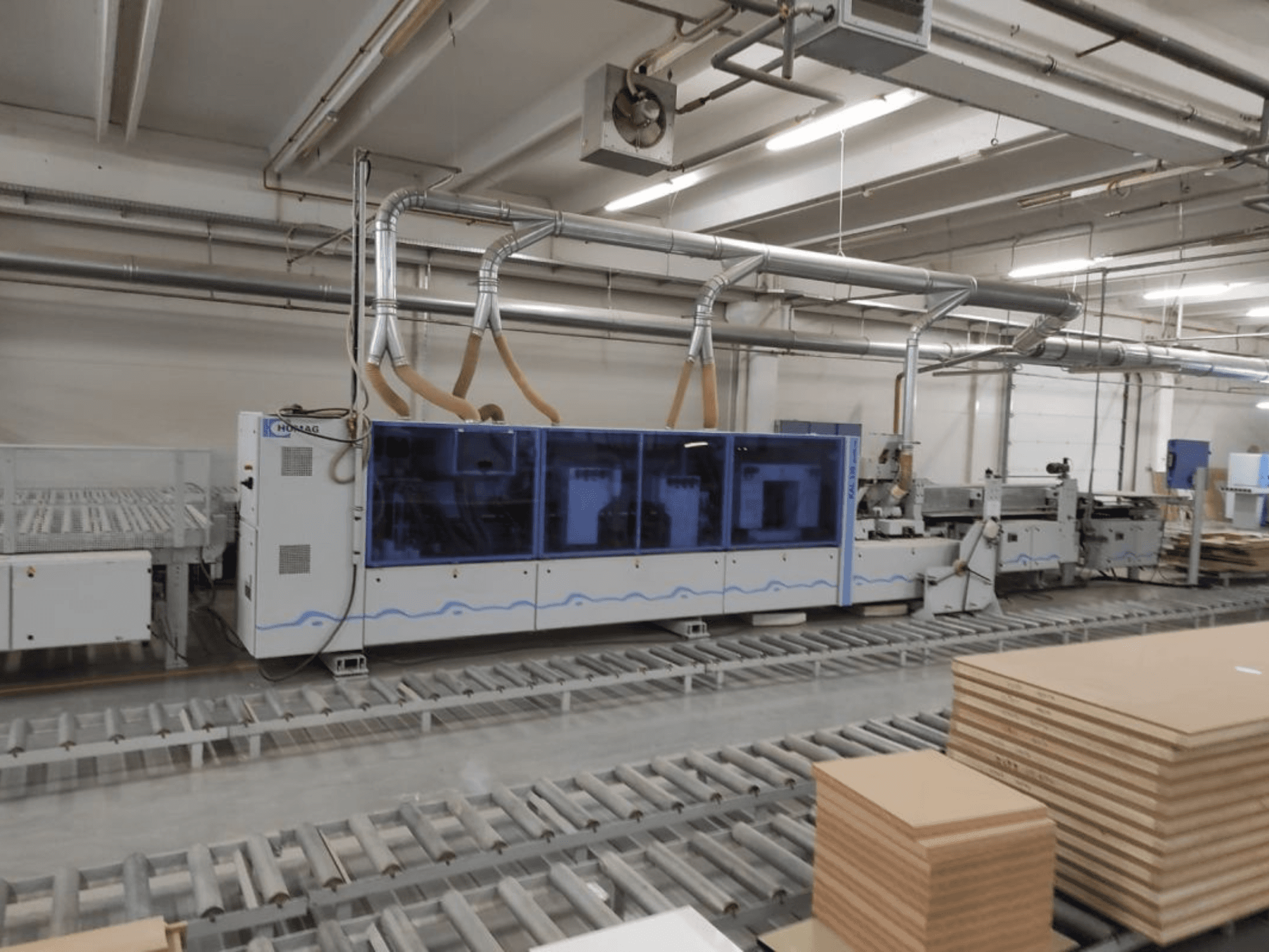 HOMAG produktionsmaskin i en fabrik, vy framifrån, med blått hölje och transportsystem med staplade plywoodskivor.