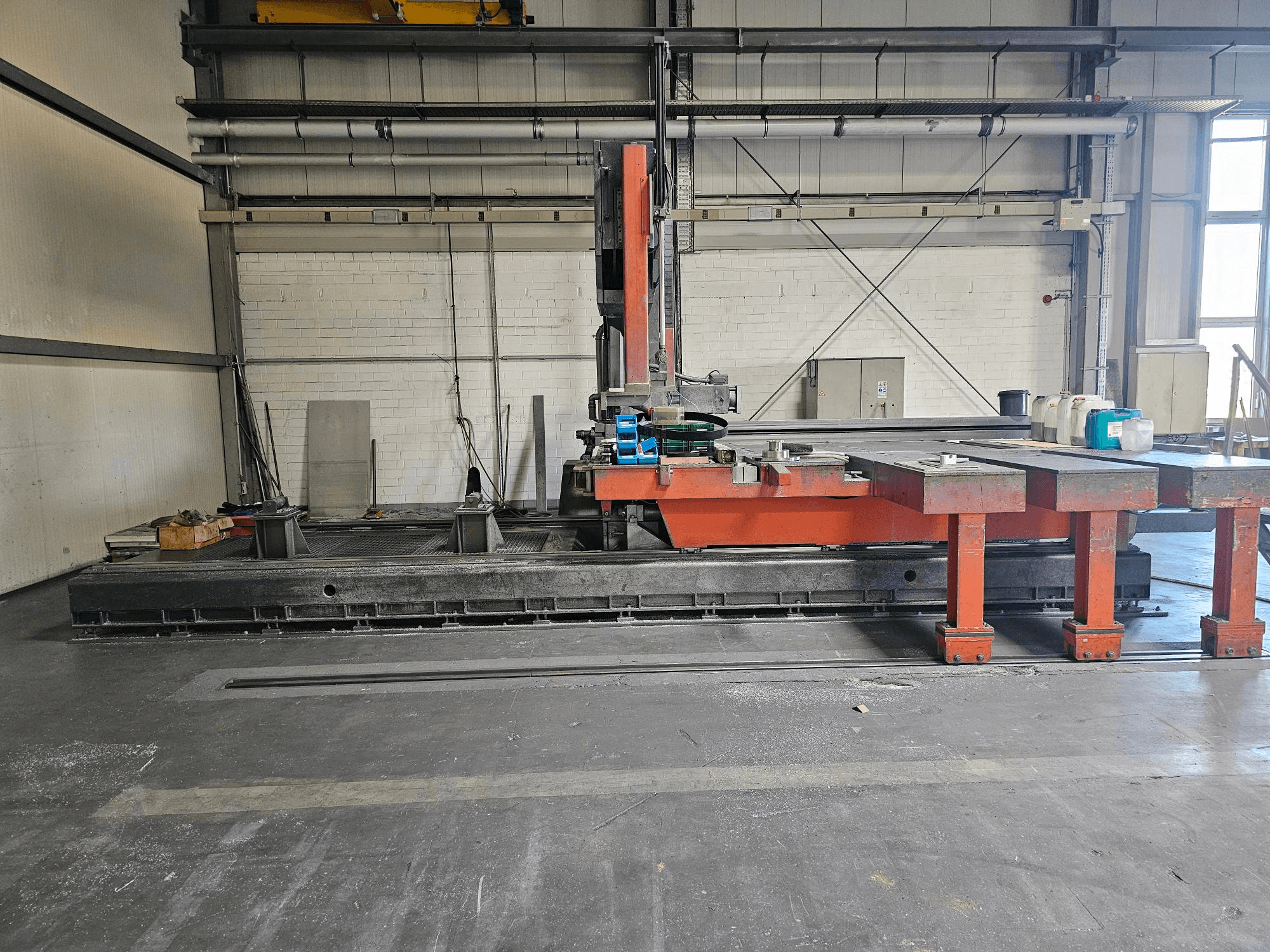 Industriell skärmaskin i en öppen verkstad, orange och svart med ett stort bord; sidovy som visar tillbehör och arbetsyta.