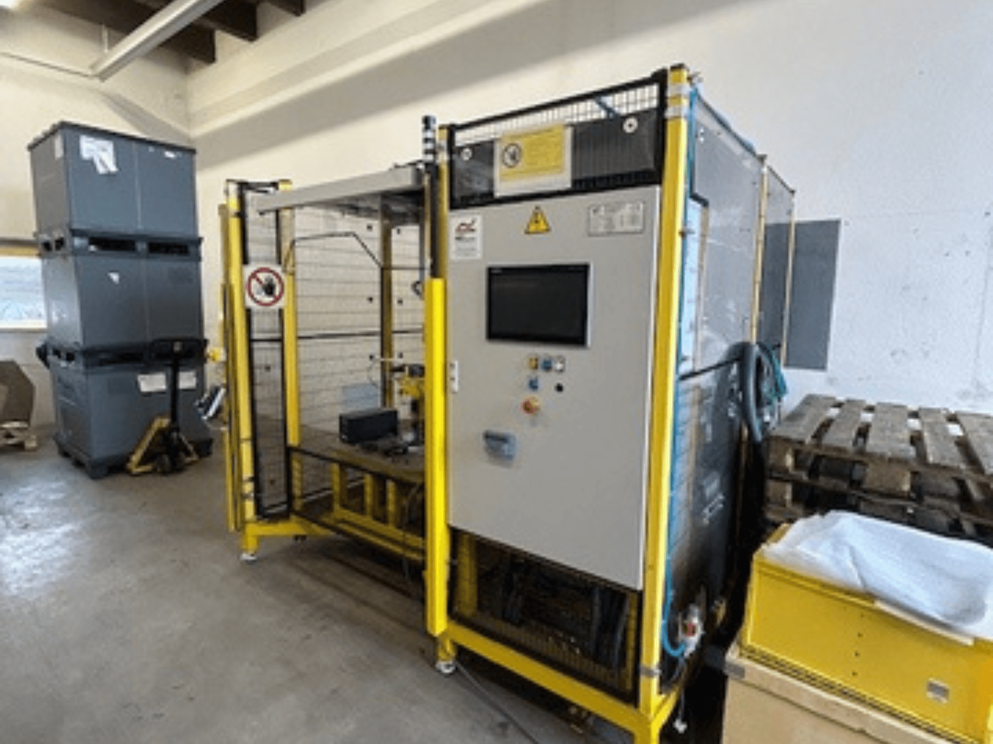 Industrimaskin med kontrollpanel och säkerhetsanordningar, framifrån; gul ram, gråa fack och pallar i bakgrunden.