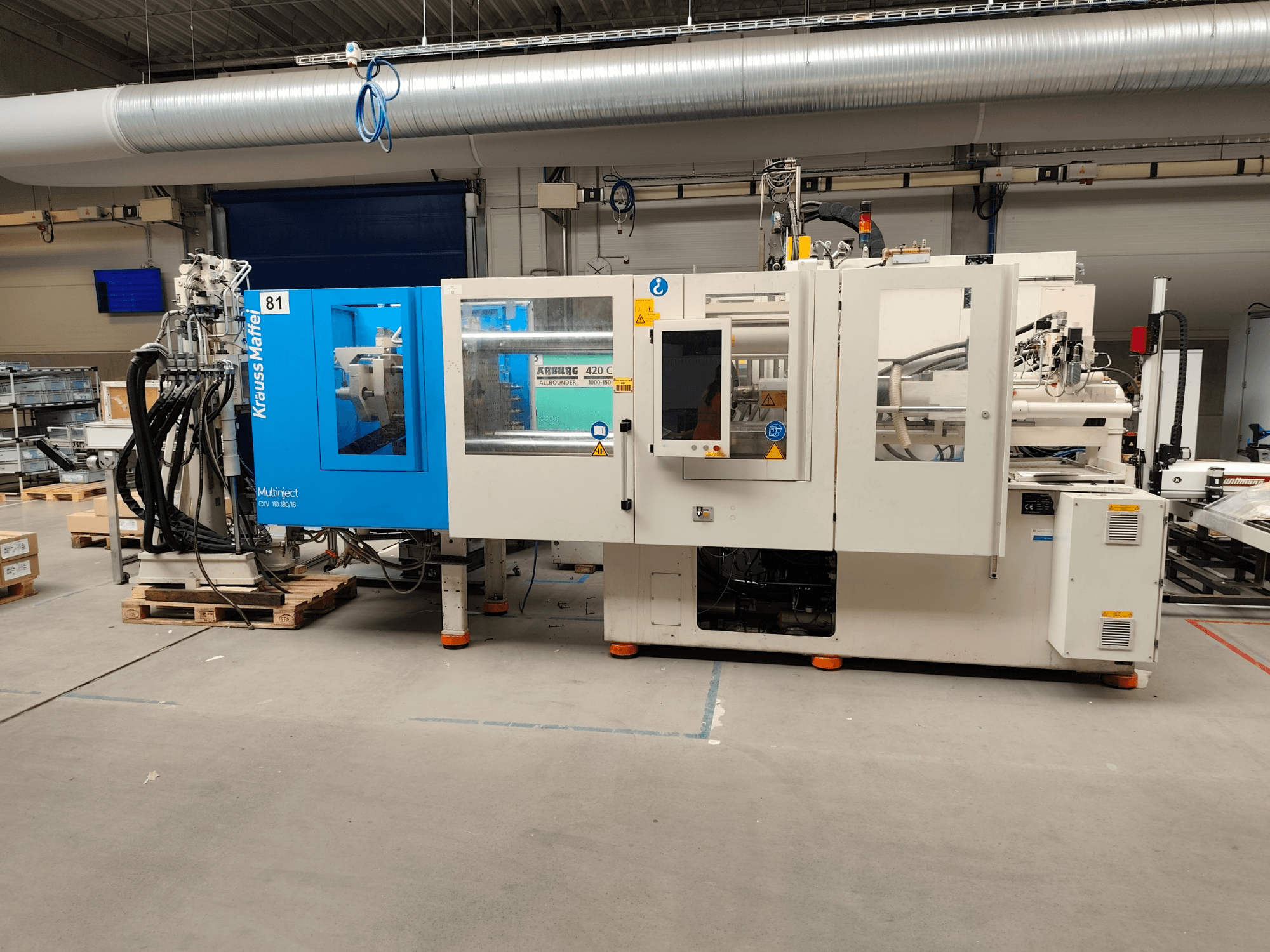 Krauss Maffei 110-180-180 CX V hydraulisk formsprutningsmaskin, sidovy, som visar den blå och vita maskinkroppen, kontrollpanelen och anslutningarna.
