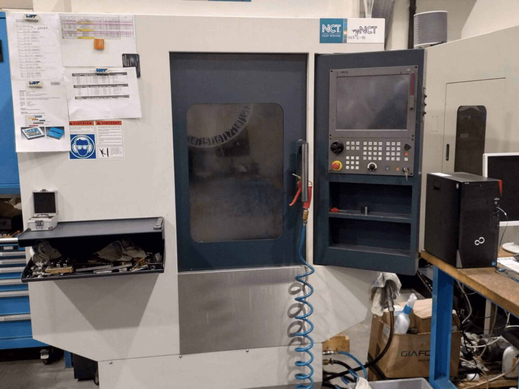 NCT CNC-maskin, vy framifrån, med kontrollpanel med numeriskt tangentbord, verktyg i förvaringslåda och manual på väggen.