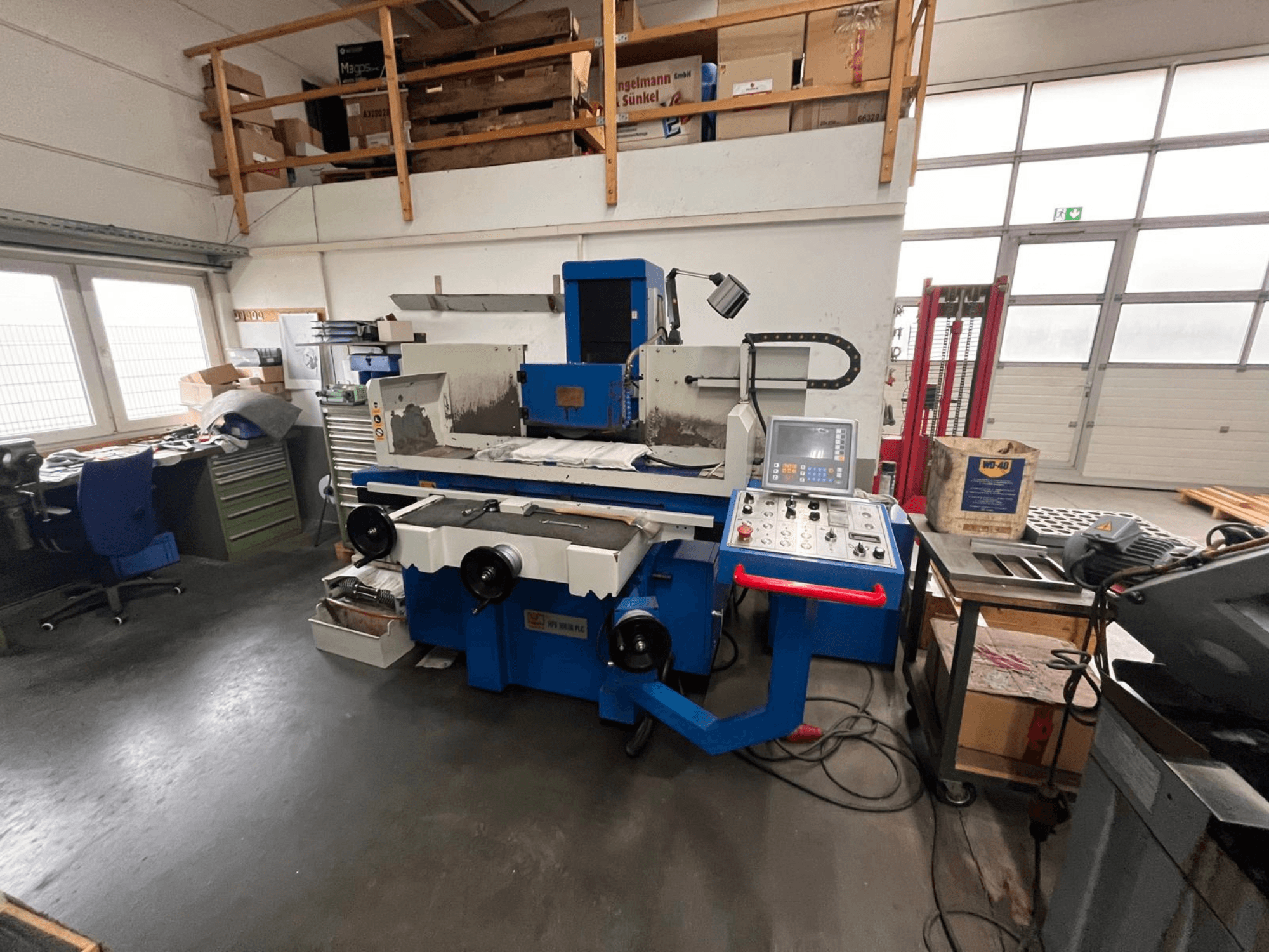 Blå planslipmaskin med digital kontrollpanel, vy framifrån, med rörigt arbetsutrymme och ett grönt verktygsskåp.