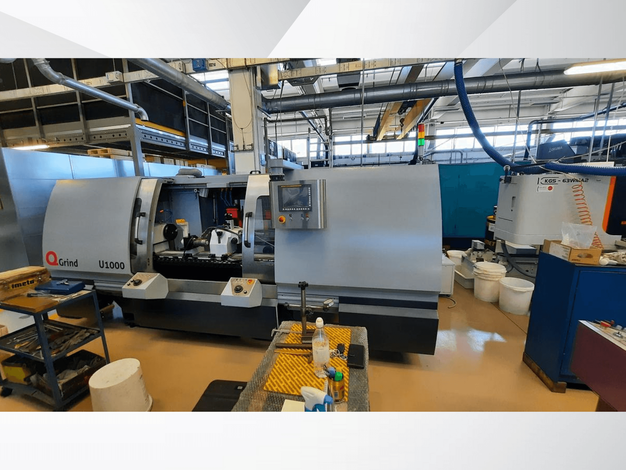 Q-Grind U1000-maskin i en verkstad, som visar en frontvy med kontrollpanel och öppet skärområde, omgiven av utrustning.