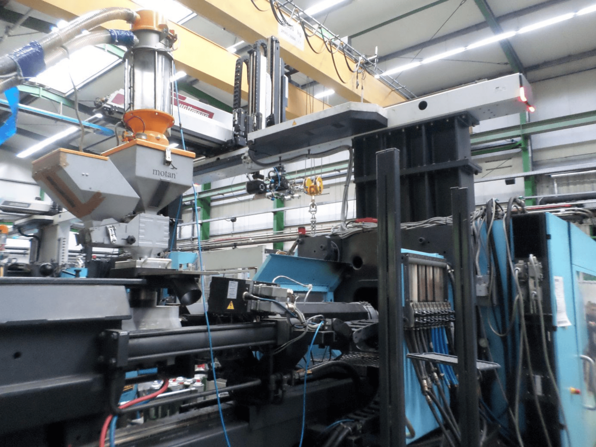 Hydraulisk formsprutningsmaskin DEMAG D 1300, sidovy, som visar matartunnan, styrenheten och anslutningarna i en fabriksmiljö.