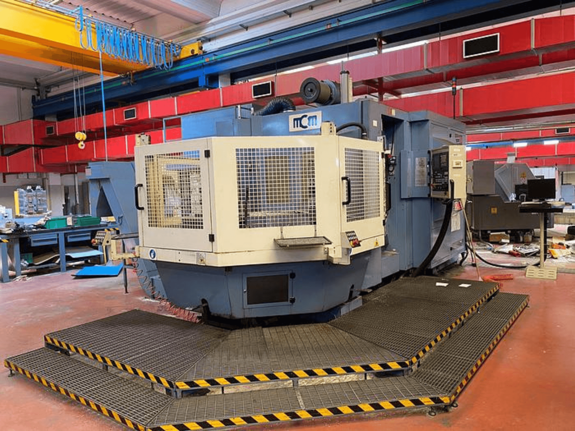 MCM CNC-maskin i en fabrik, framifrån med skyddsbur, svarta och gula säkerhetslister, omgiven av industriutrustning.