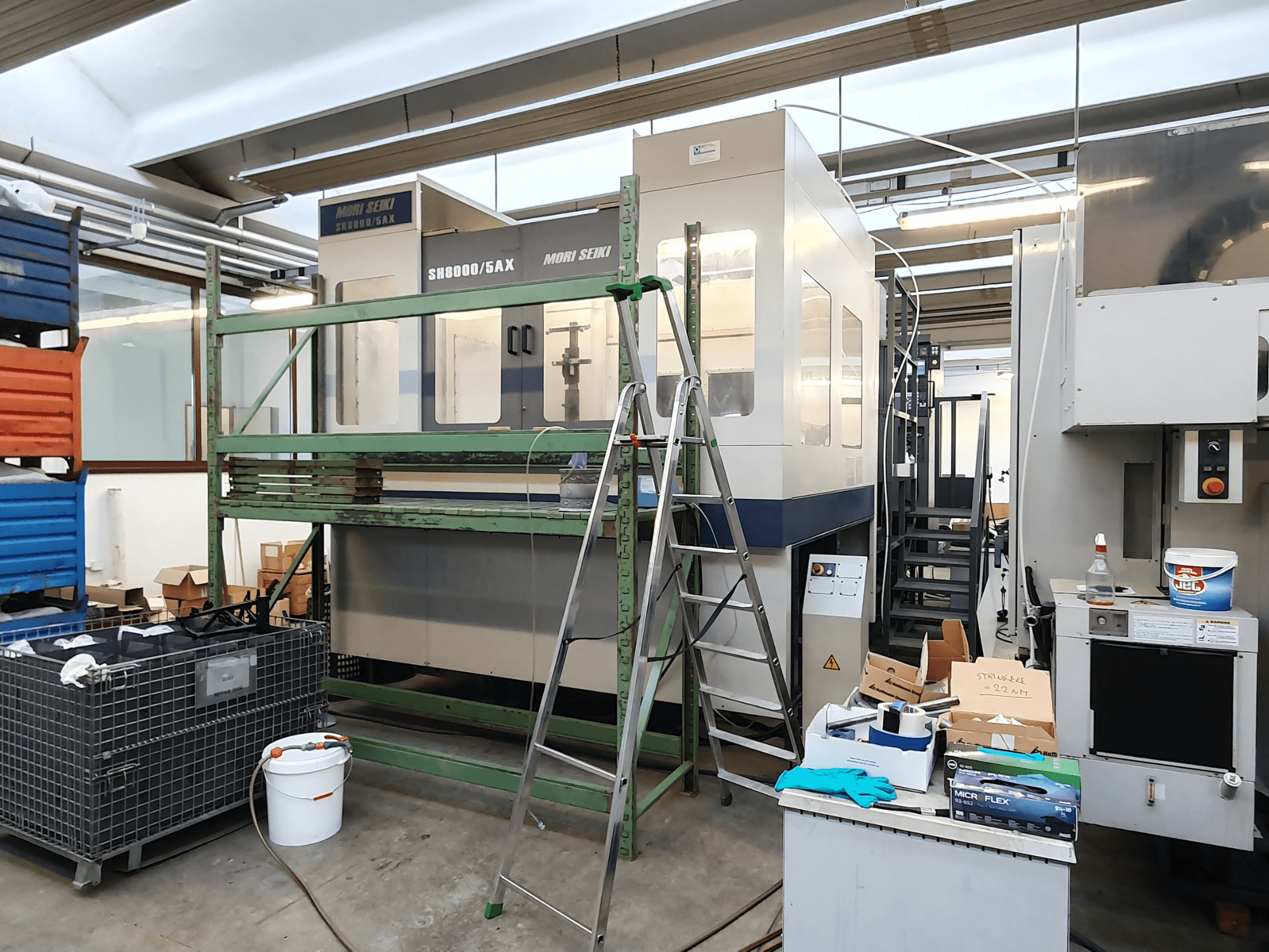 Mori Seiki SH-8000/5AX CNC-maskin i en verkstad, vy framifrån, med verkstadsverktyg och förvaringslådor i närheten.