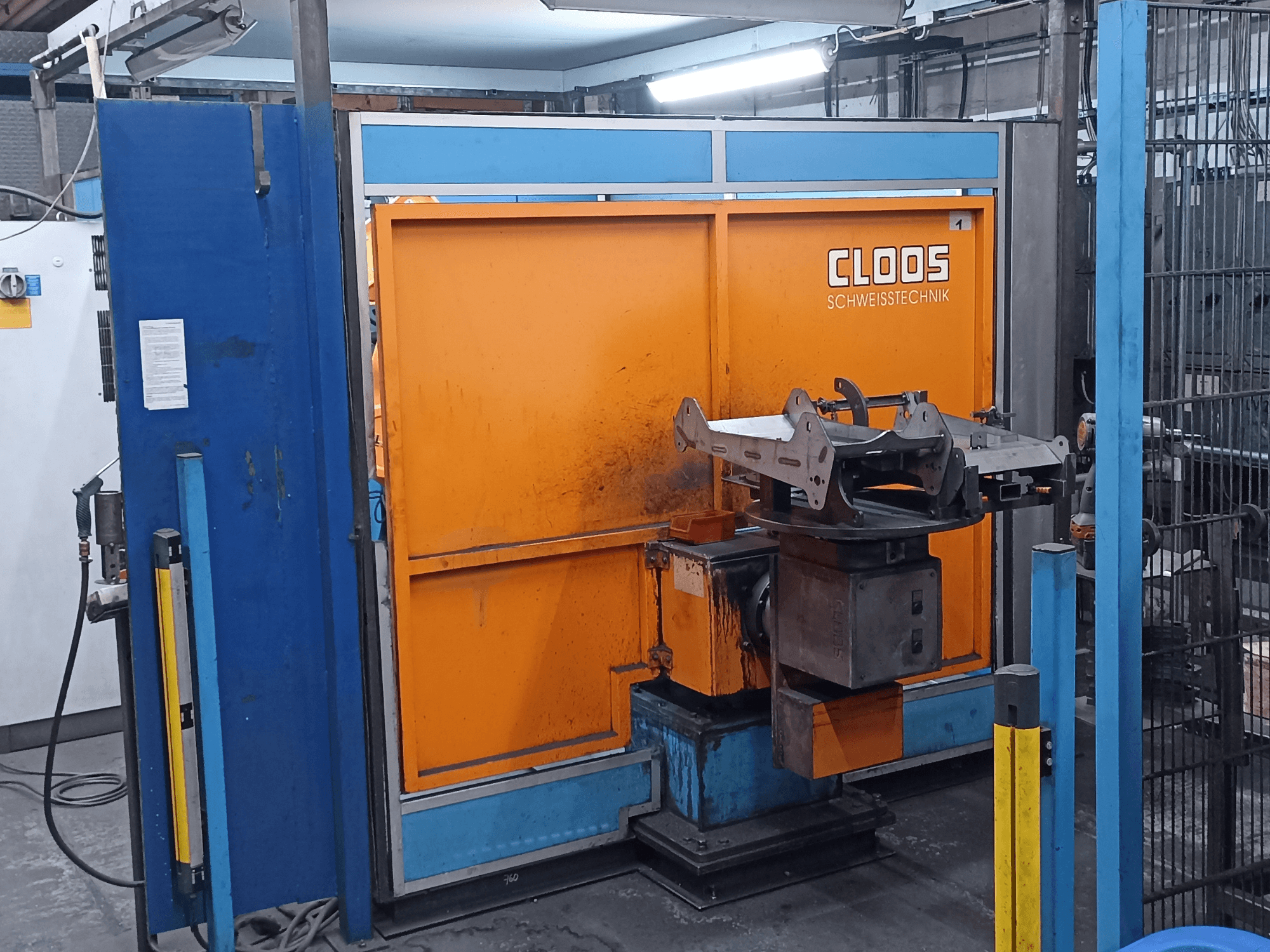 En CLOOS Romat 320-svetsrobot i en verkstad, sett framifrån, med en orange kontrollpanel och ett arbetsstycke av metall på en roterande bas.