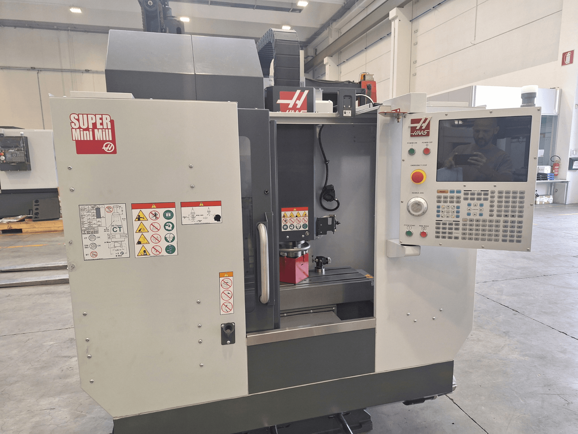 HAAS Super Mini Mill sett framifrån, med kontrollpanelen, säkerhetsetiketterna och bearbetningsområdet där ett arbetsstycke är monterat.