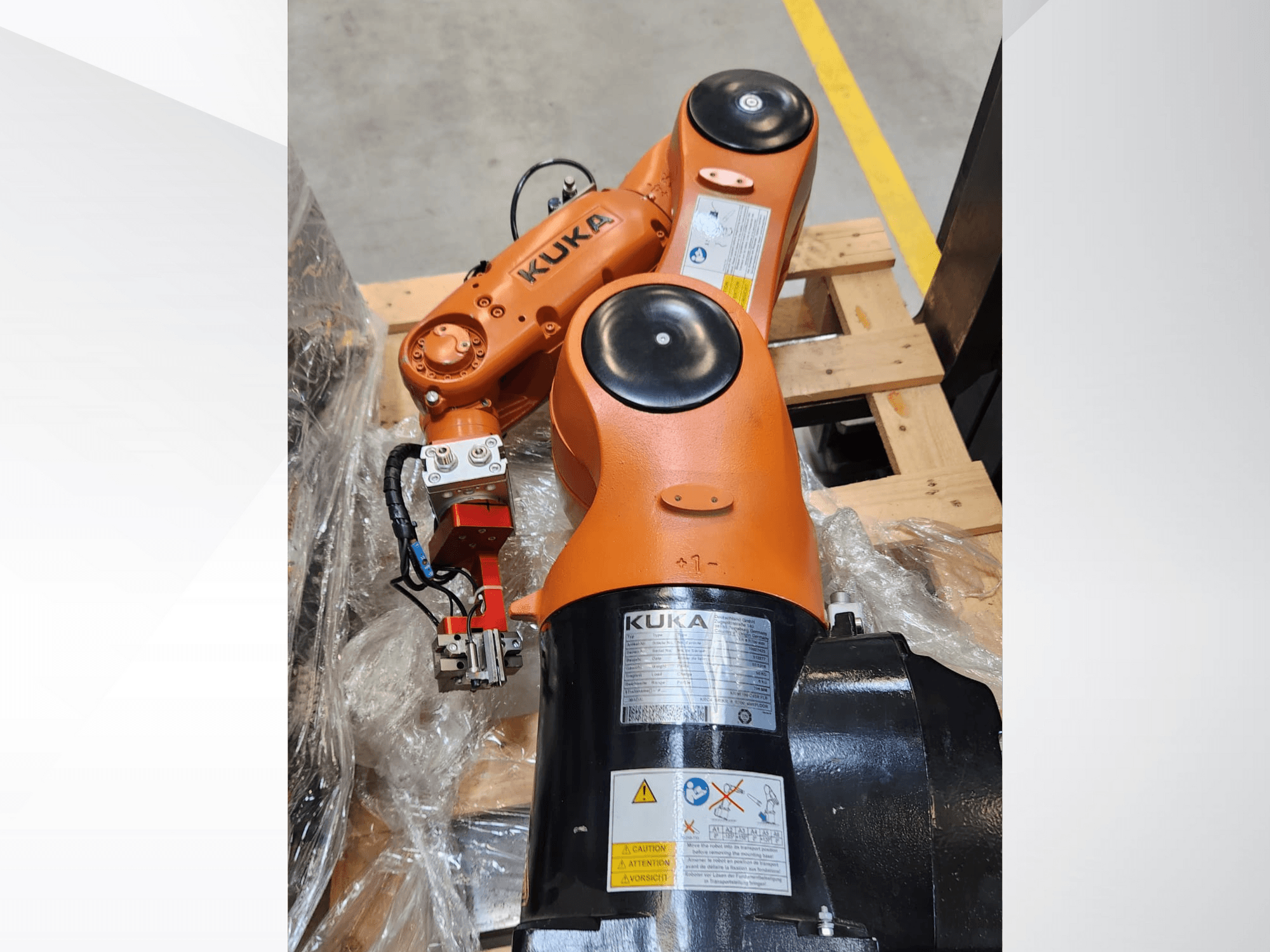 KUKA robotarm med orange finish, vy från sidan, med gripdon och styrkablar på en träpall.