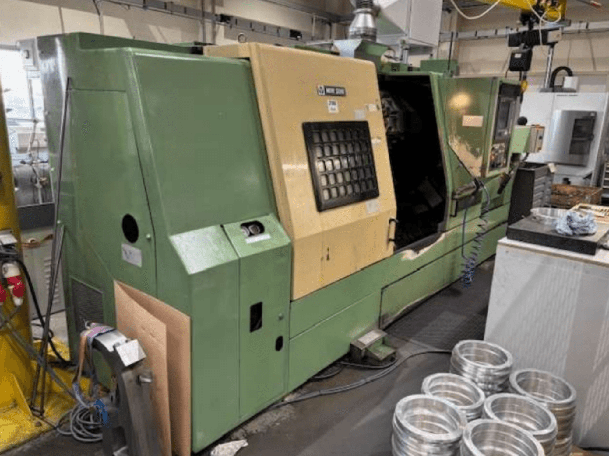 Grön CNC-svarvmaskin från Mazak, sedd framifrån, med kontrollpanel och bearbetade aluminiumdelar på ett arbetsbord.