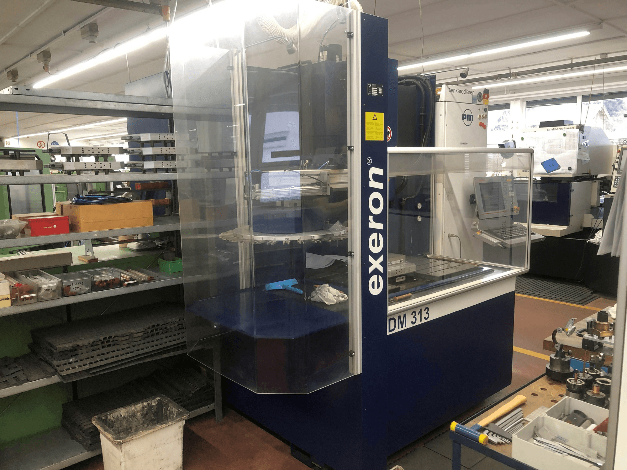 Exeron EDM 313 pressgjutningsmaskin i en verkstad, framifrån med genomskinligt hölje och kontrollpanel.