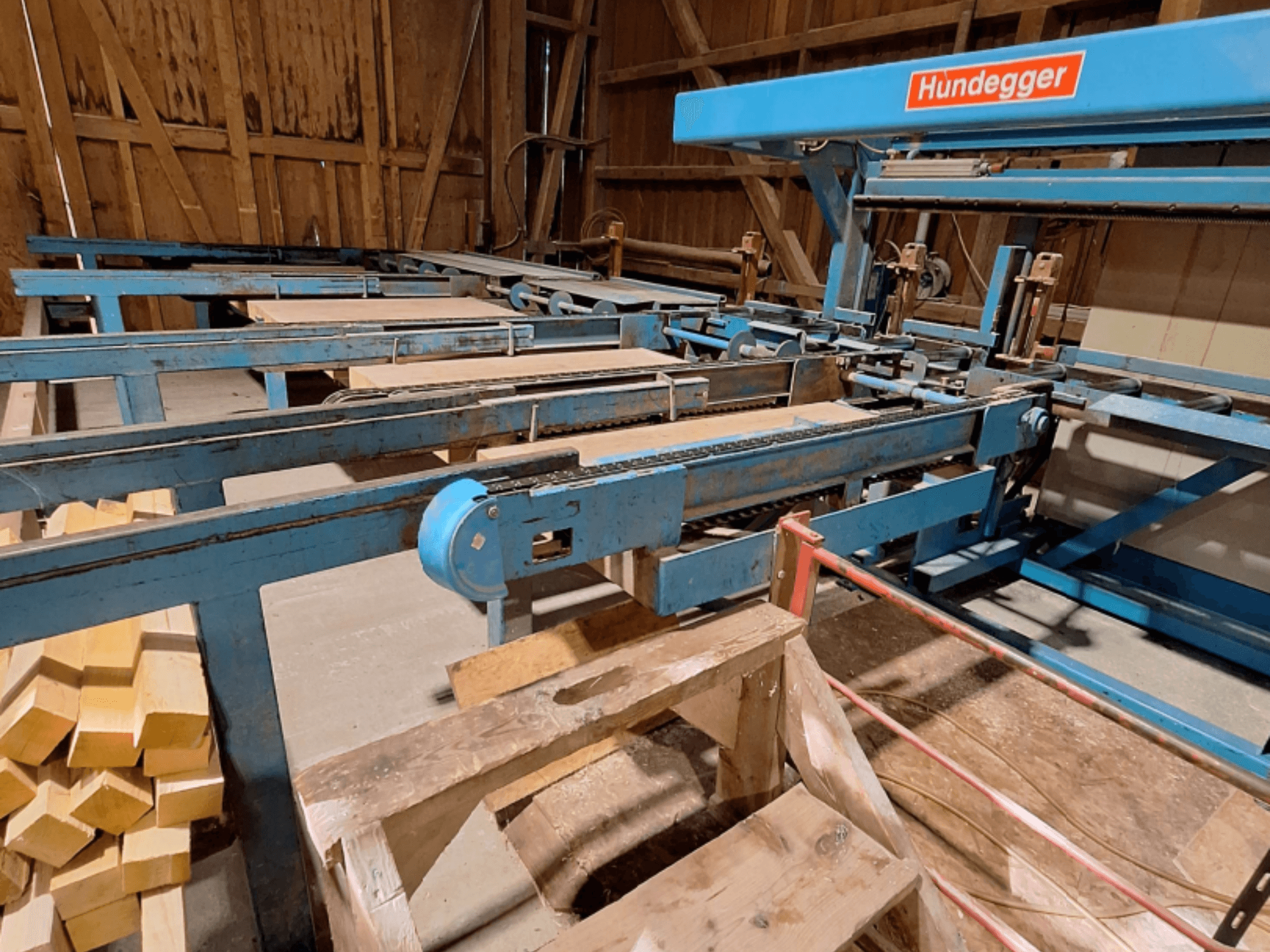 CNC-bearbetningscenter för trä HUNDEGGER K2-A i en ladugård, med blå metallram, transportörsystem och staplade träbjälkar.