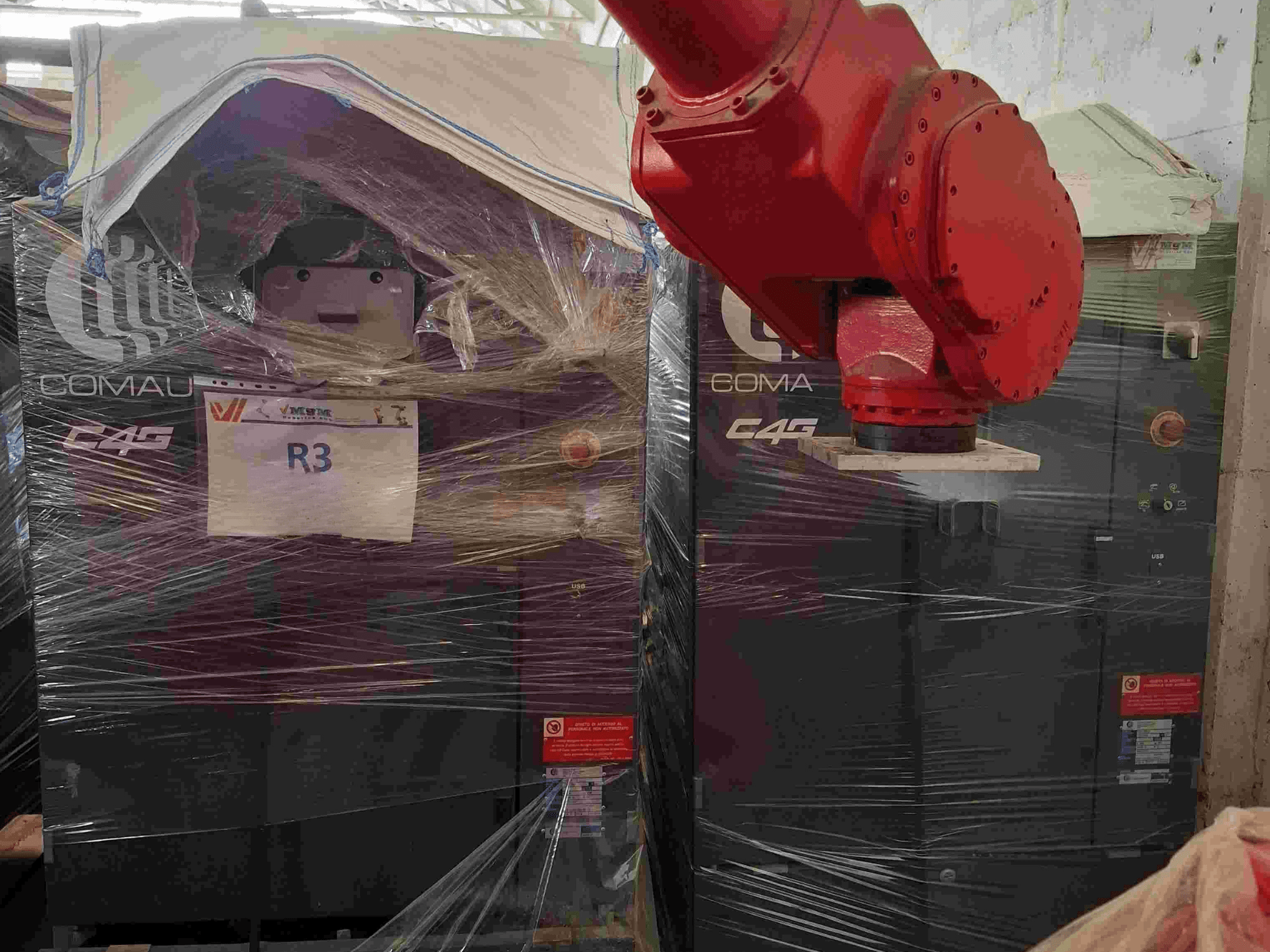 Comau C4G robotarm sedd framifrån, inslagen i plast, märkt "R3", med varningsdekaler synliga på maskinen.