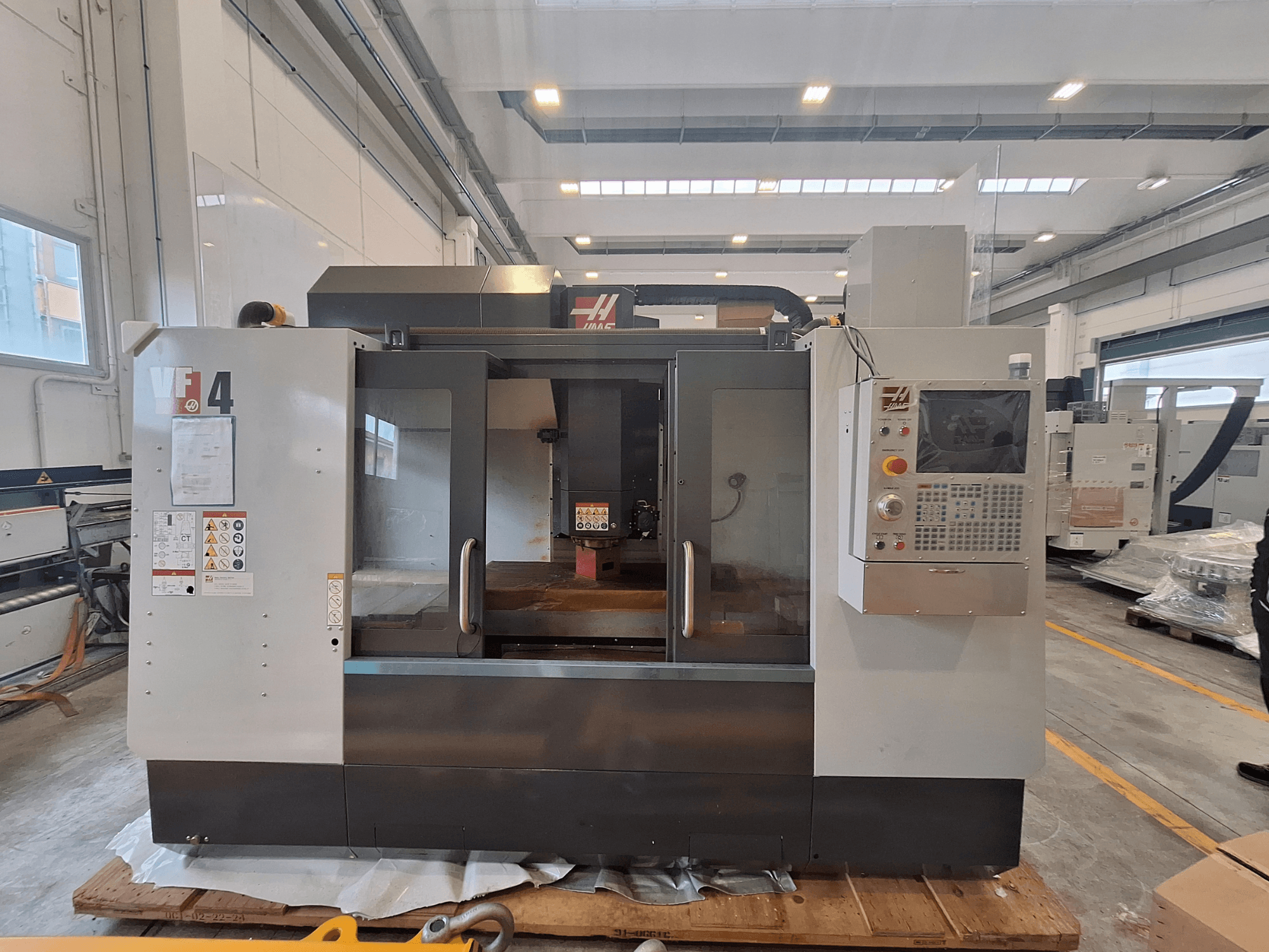 HAAS VF-4 vertikalt bearbetningscenter, framifrån, där kontrollpanelen, arbetsområdet och säkerhetsmärkningar syns i en lagermiljö.
