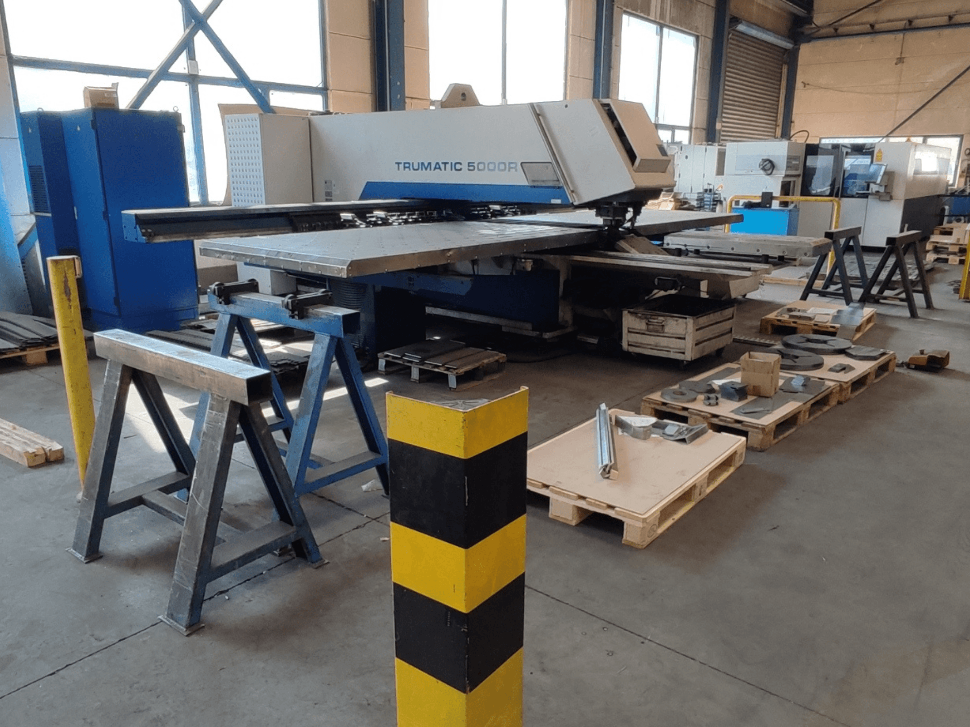 En TRUMPF Trumatic 5000 R CNC-stansmaskin i en verkstad, sett från sidan med verktyg och material på pallar i närheten.