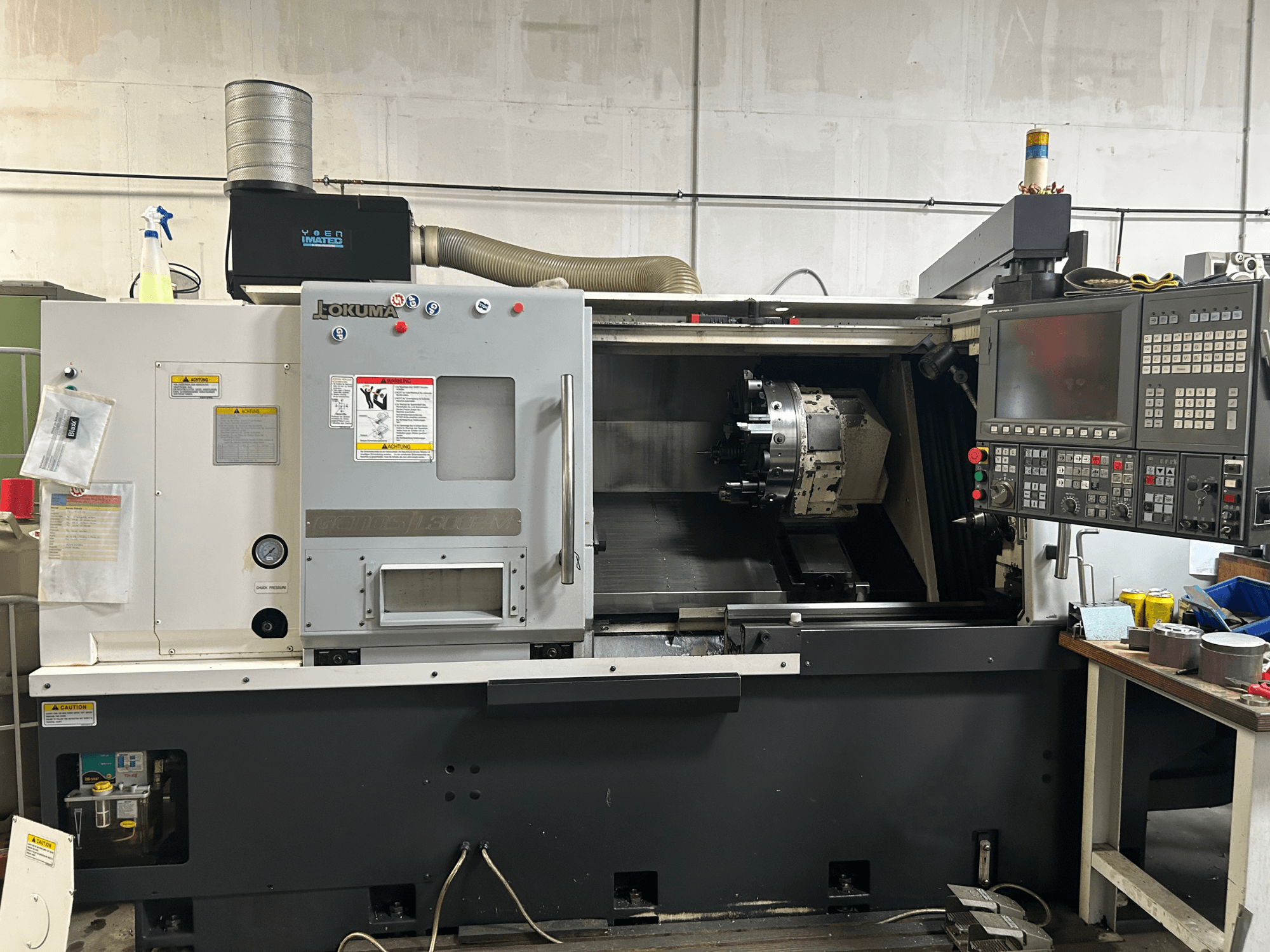 Frontvy av en Okuma Genos L300E-M horisontell svarv med kontrollpanel, synlig spindel och olika uppställningsverktyg.