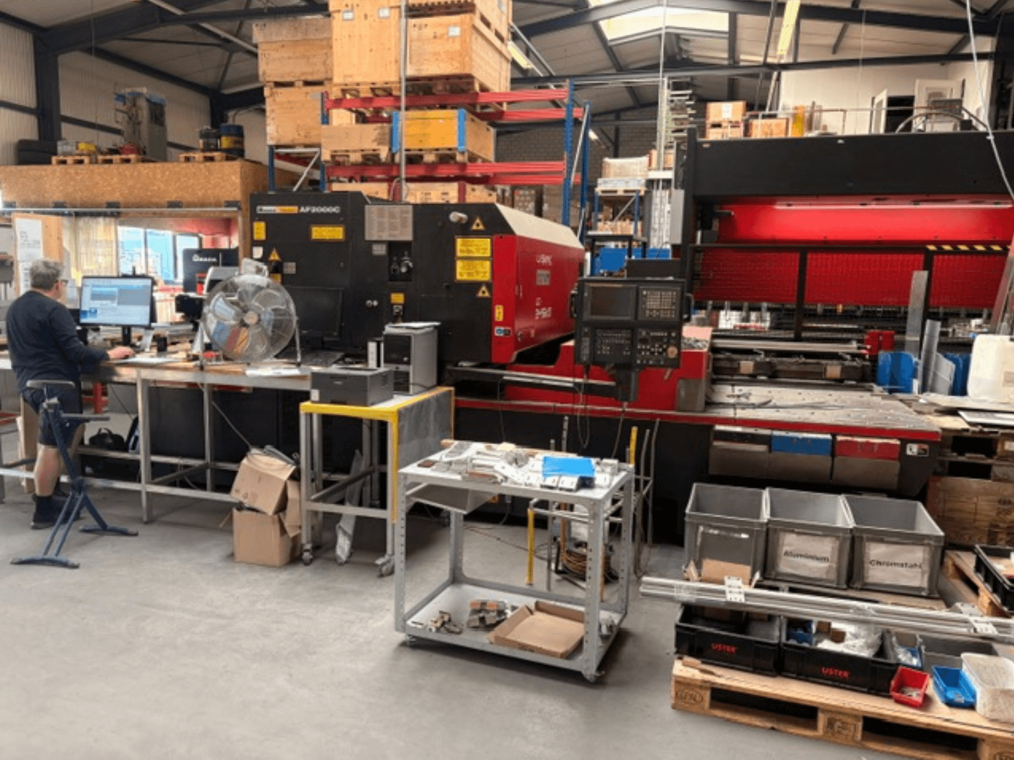 En AMADA LC-2415αIII CO₂-laserskärmaskin i en verkstadsmiljö, med en datorarbetsstation synlig i förgrunden.