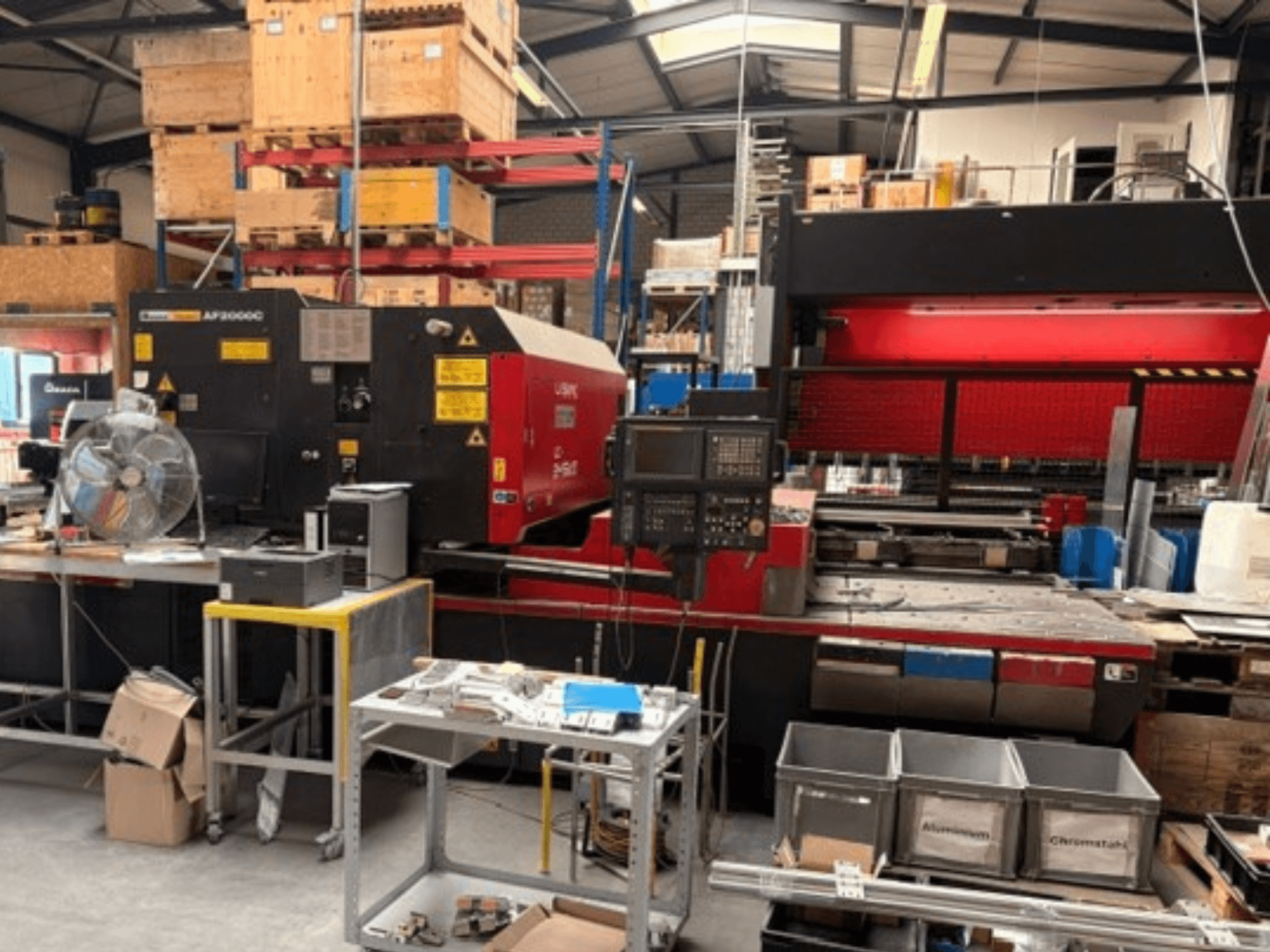 En AMADA LC-2415αIII CO₂-laserskärmaskin i en verkstadsmiljö, med framifrånvy, kontrollpanel och arbetsutrymme.