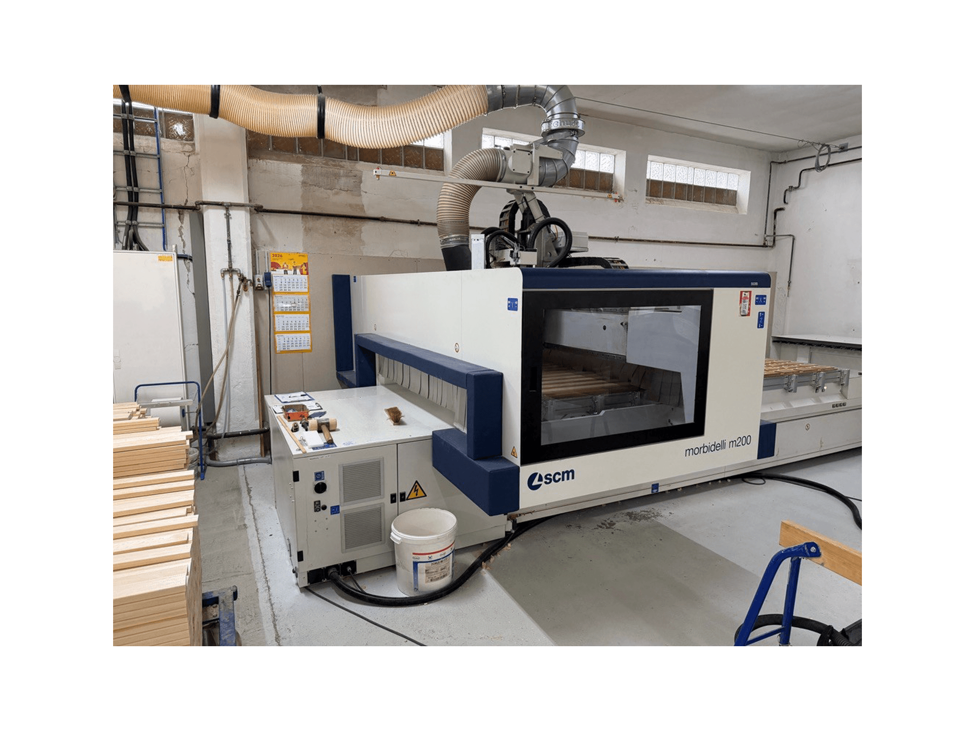 Ett SCM Morbidelli M200F CNC-bearbetningscenter i en verkstad, sett från sidan med träbitar på bordet och ett ventilationssystem ovanför.