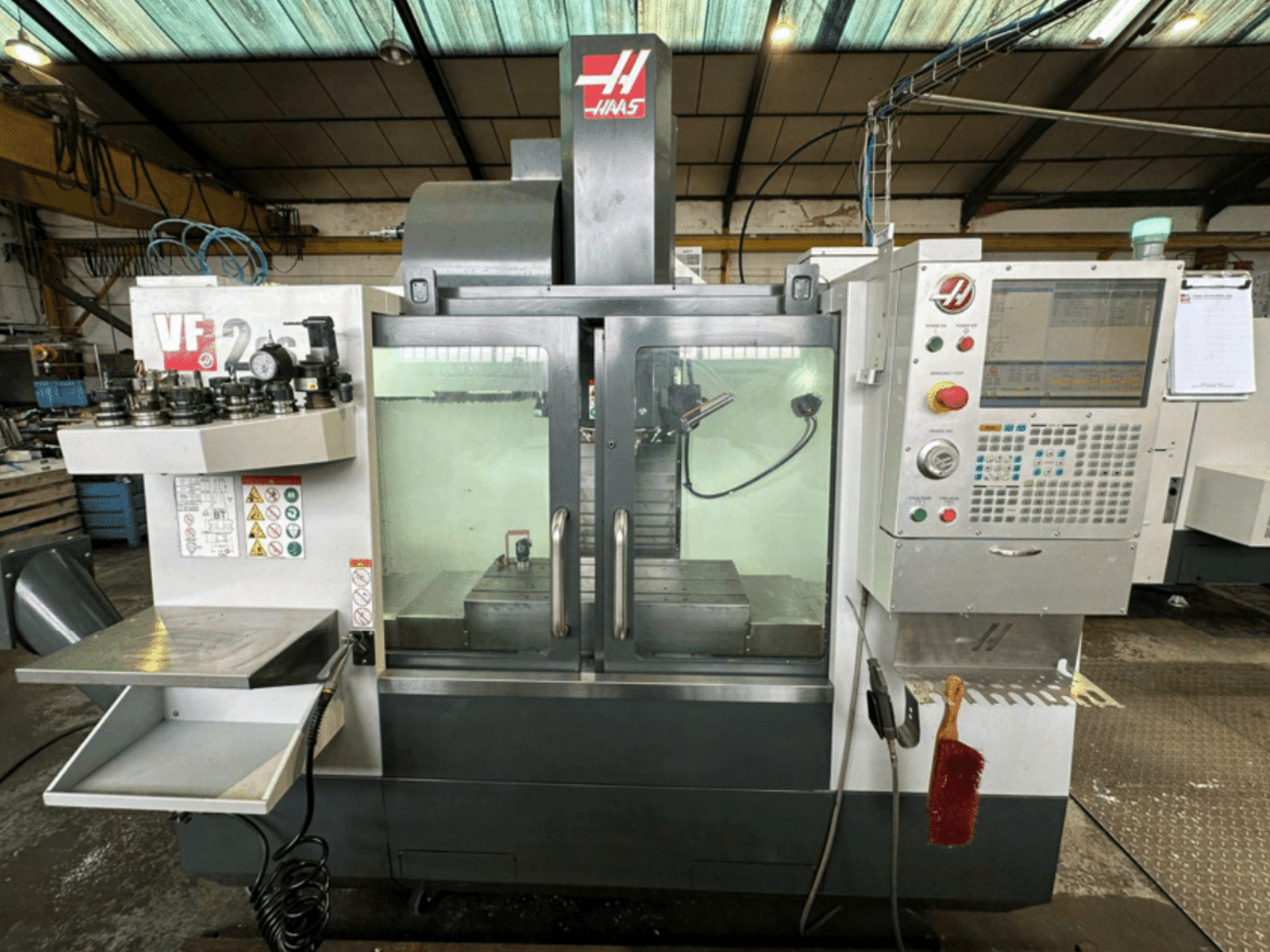 Haas VF2 CNC-maskin framifrån, med kontrollpanel, verktygshållare och arbetsyta med säkerhetsetiketter och en borste.