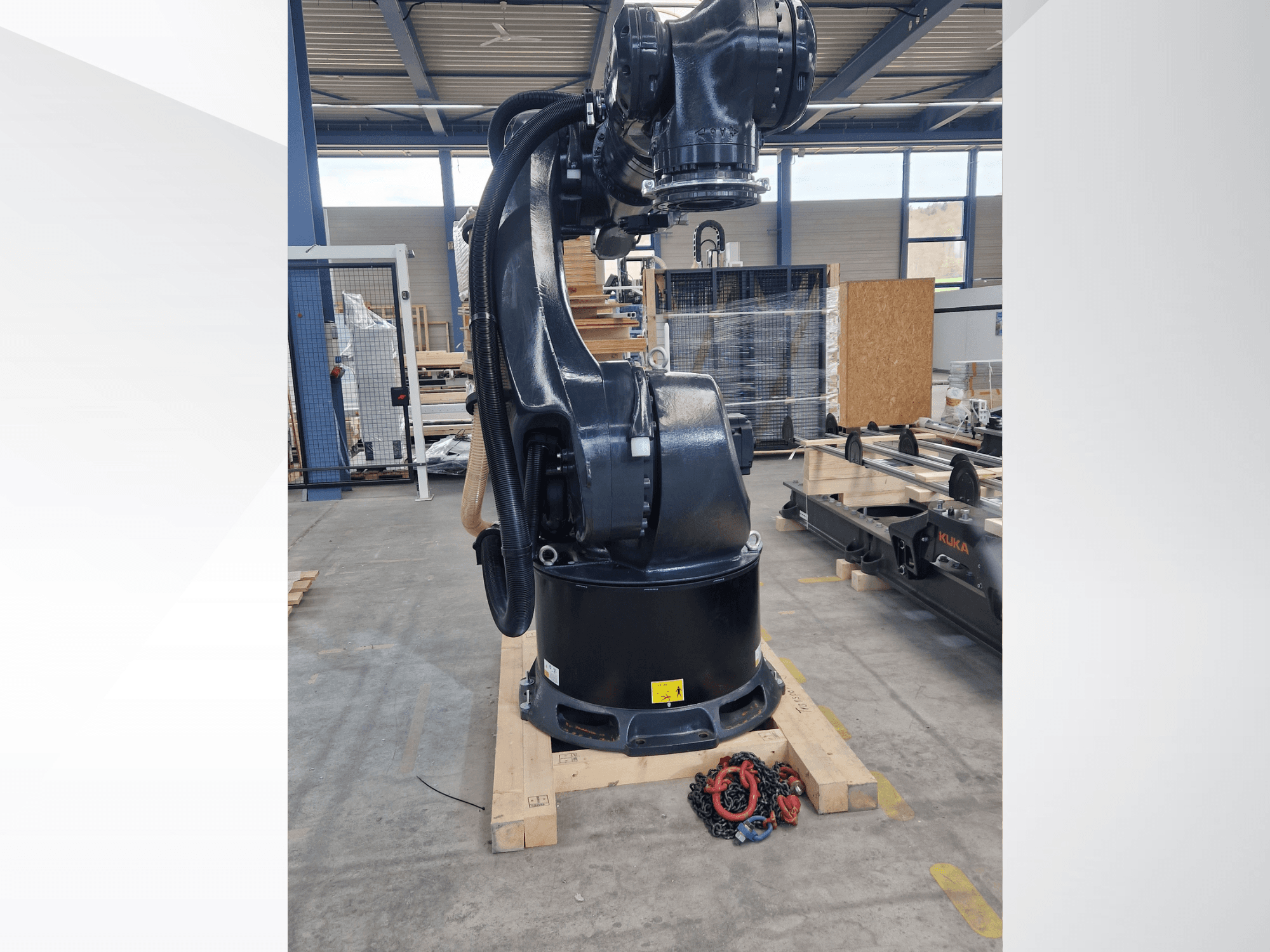 En KUKA KR 340 R3330-robotarm i ett lager, sett från sidan, där basen och kablarna syns, med kedjor i förgrunden.