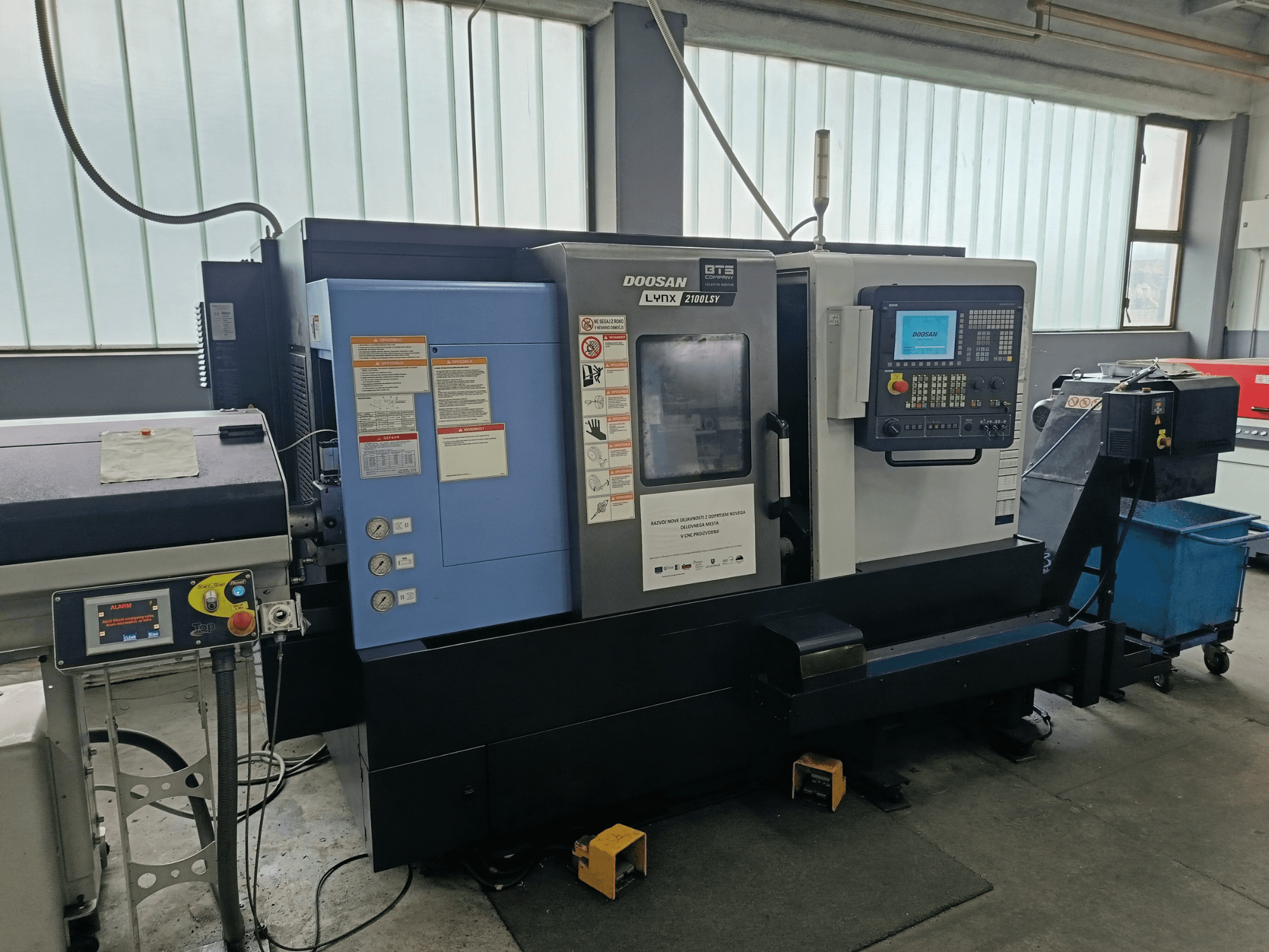 DOOSAN LYNX 2100 LSY Turn-Mill Center, sidovy som visar kontrollpanelen och manöverknappar, med informationsetiketter synliga.