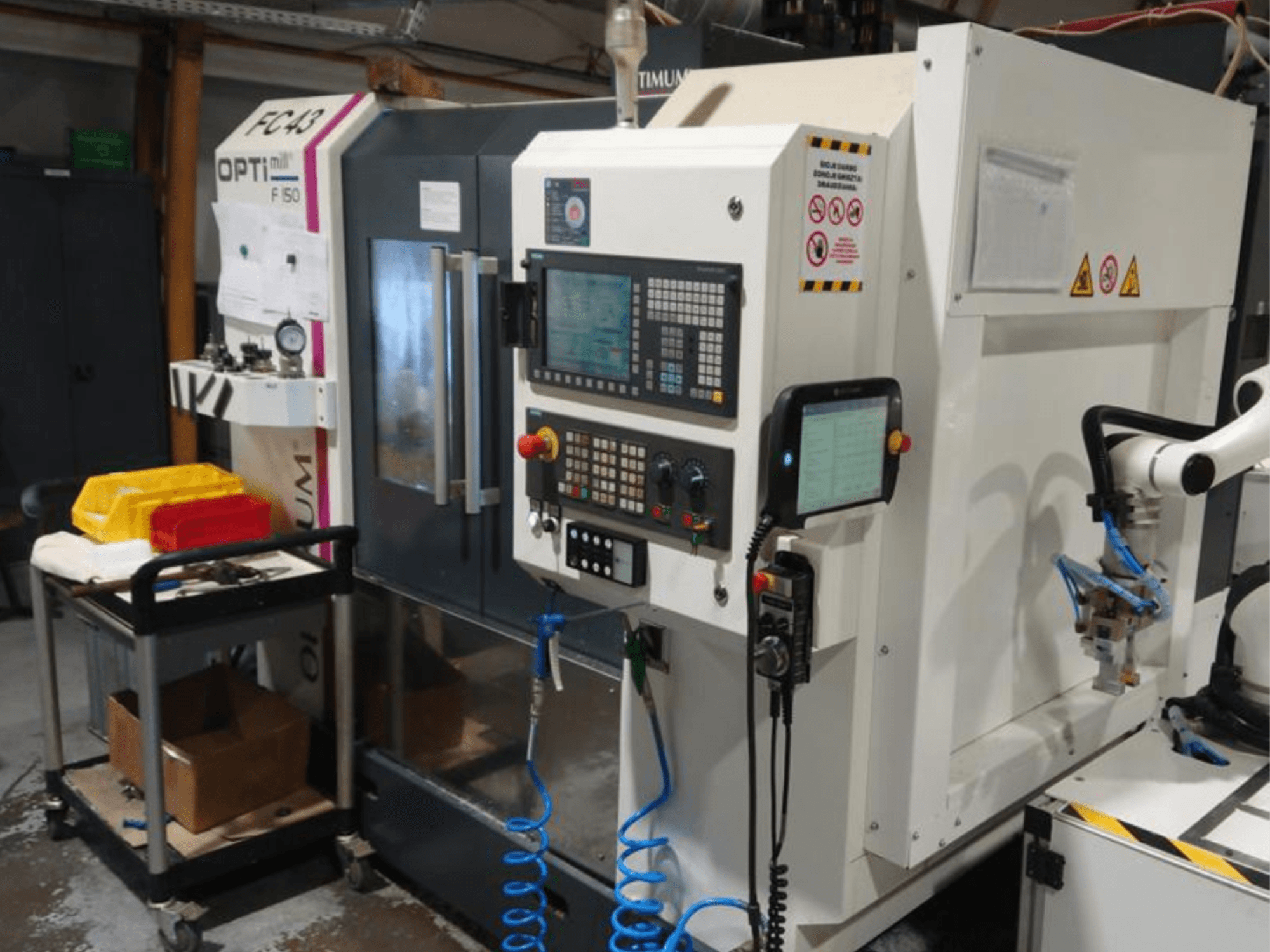 Kontrollpanelen på fräsmaskinen OPTIMUM F50 med slangar, verktyg och en robotarm i en verkstadsmiljö.