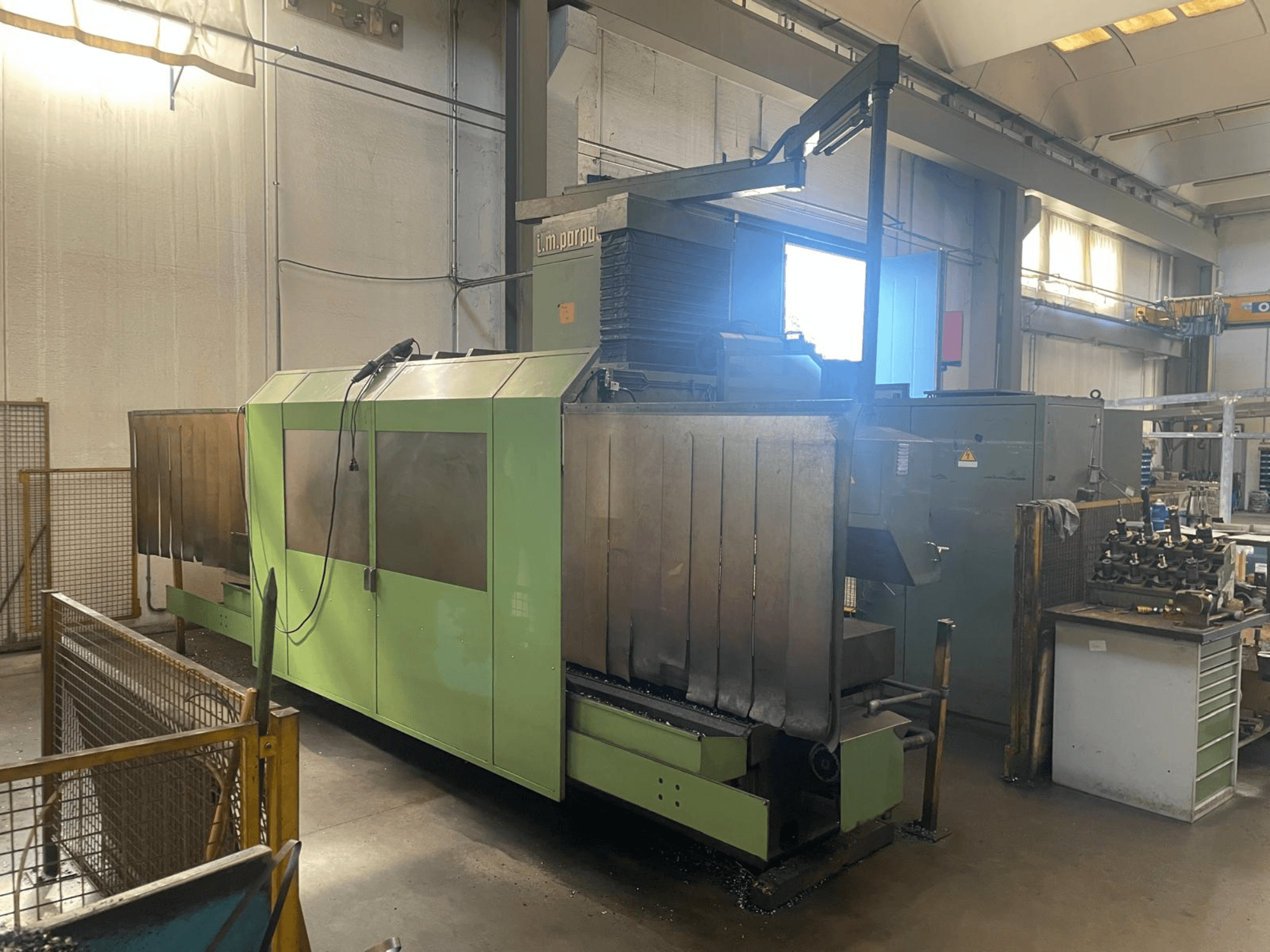 Grön industrimaskin med synliga metallkomponenter, framifrån; har kontrollpanel, säkerhetsvarningar och arbetsstation i närheten.