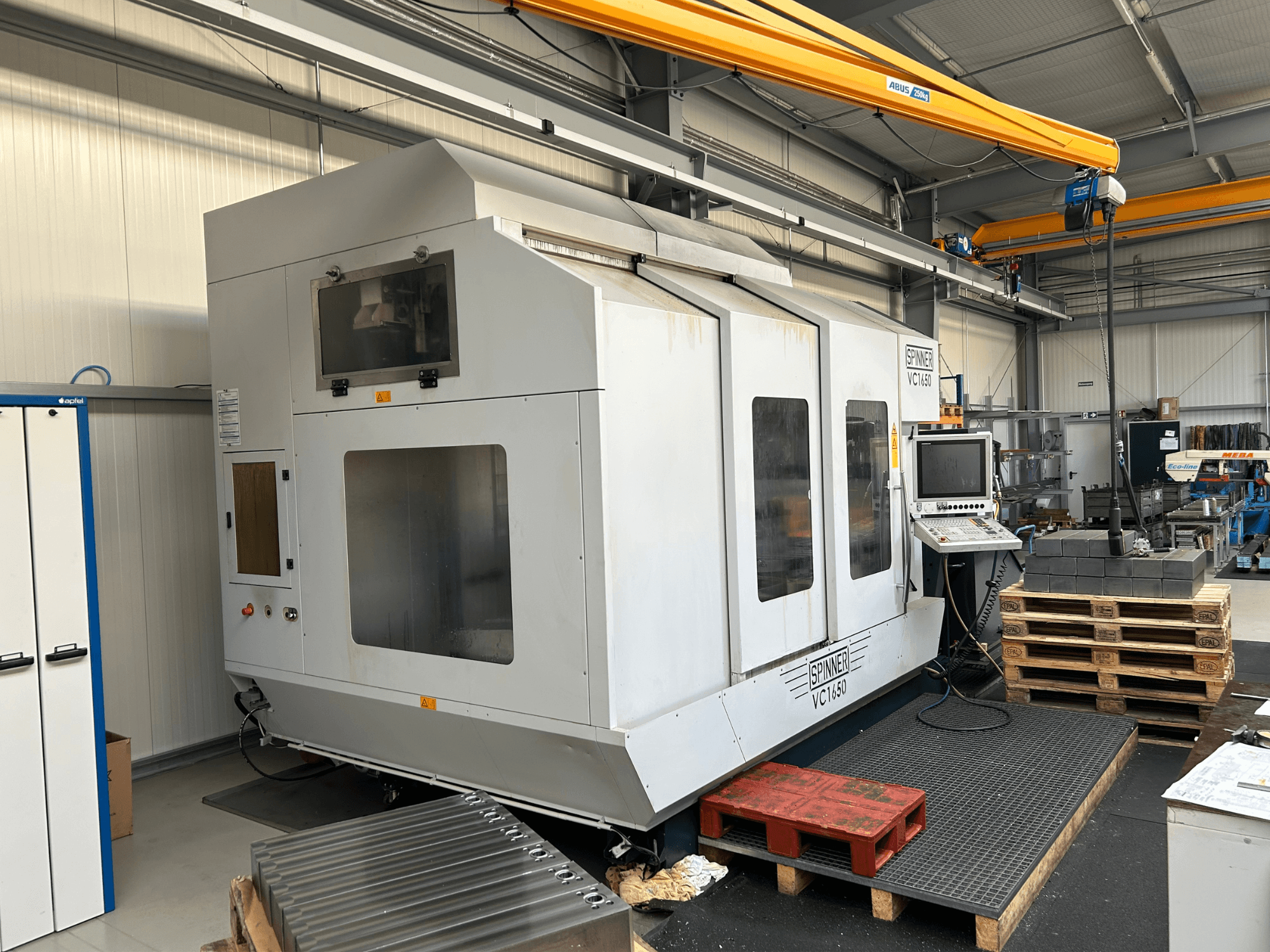 Spinner VC 1650 CNC-maskin, framifrån, med kontrollpanel, stora fönster och pallar staplade i närheten i en verkstadsmiljö.