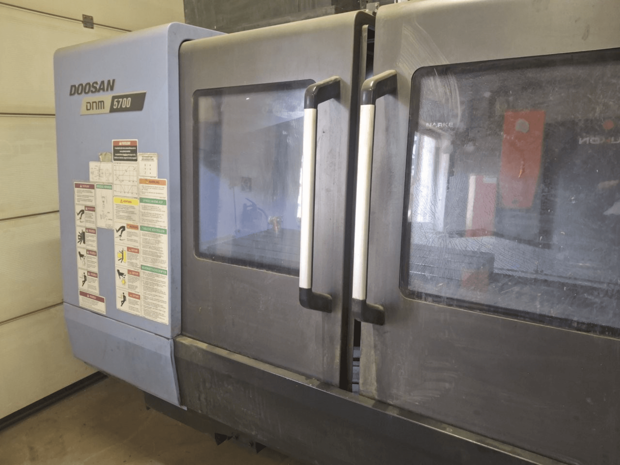 Frontvy av en DOOSAN DNM 5700 vertikal fleroperationsmaskin med dubbla dörrar och kontrollpaneler med riktlinjer för driften.