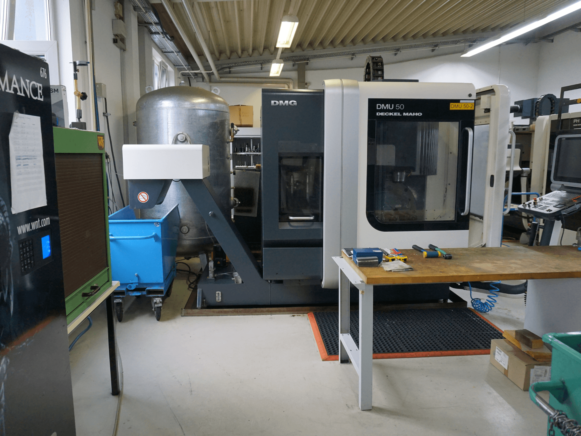 DMG DMU 50 fräsmaskin i en verkstad, framifrån, med verktyg på ett träbord och en blå soptunna med hjul i närheten.