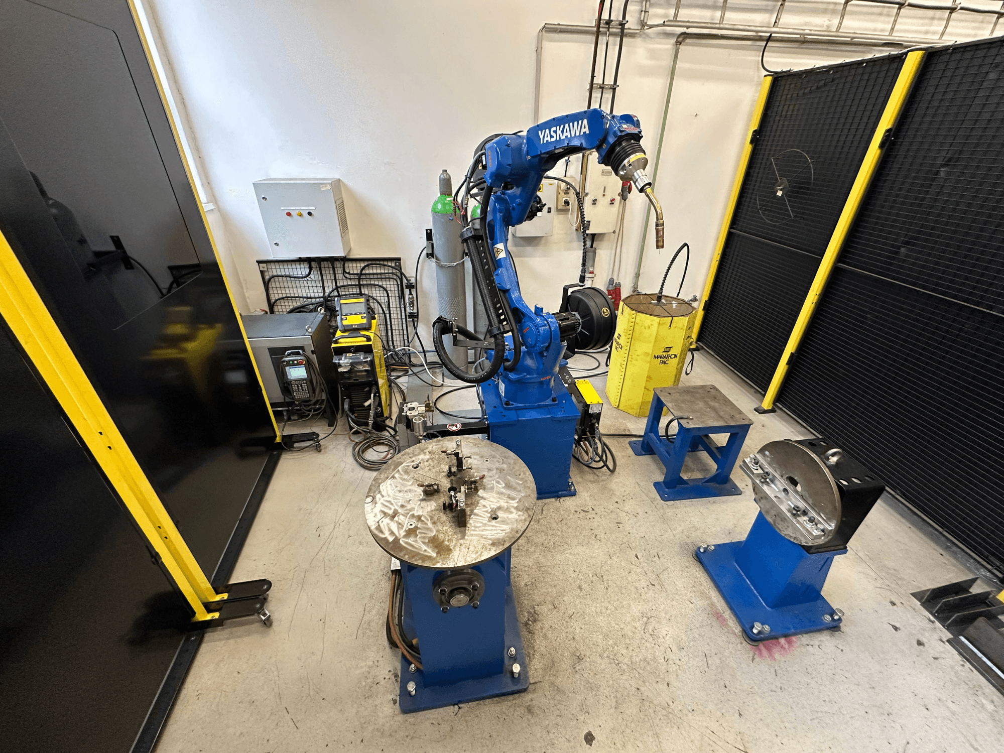 Yaskawa robotarm i blått med två arbetsbord, svetsutrustning och säkerhetsbarriärer; industriellt arbetsutrymme sett framifrån.