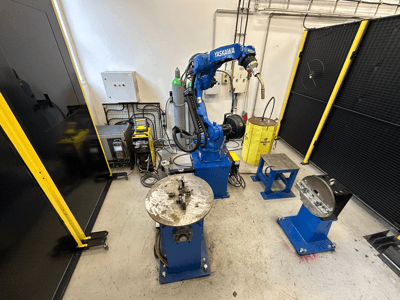 Yaskawa robotarm i blått med två arbetsbord, svetsutrustning och säkerhetsbarriärer; industriellt arbetsutrymme sett framifrån.