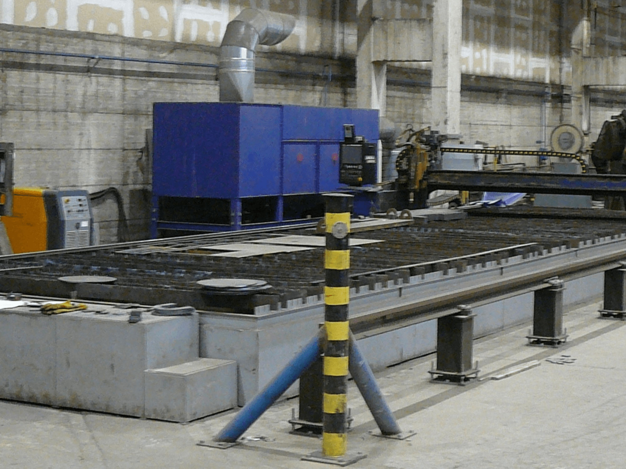 Uppställning av industriell metallskärmaskin, vy från sidan, med blå kontrollbox, säkerhetsremsor och arbetsyta i metall.