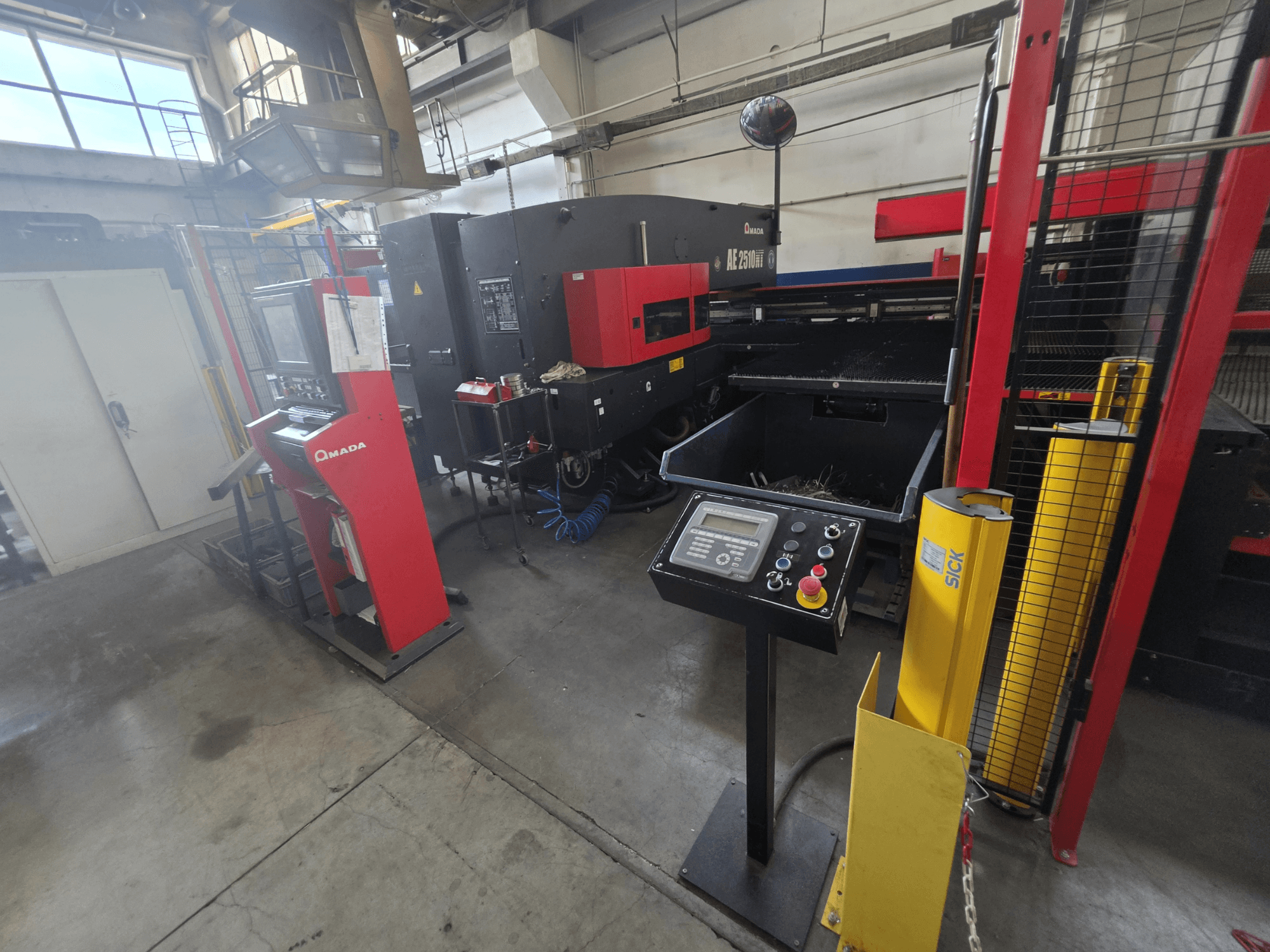 CNC-stansmaskin AMADA AE-2510NT, framifrån med kontrollpanel, säkerhetsfunktioner och den omgivande industriella miljön.