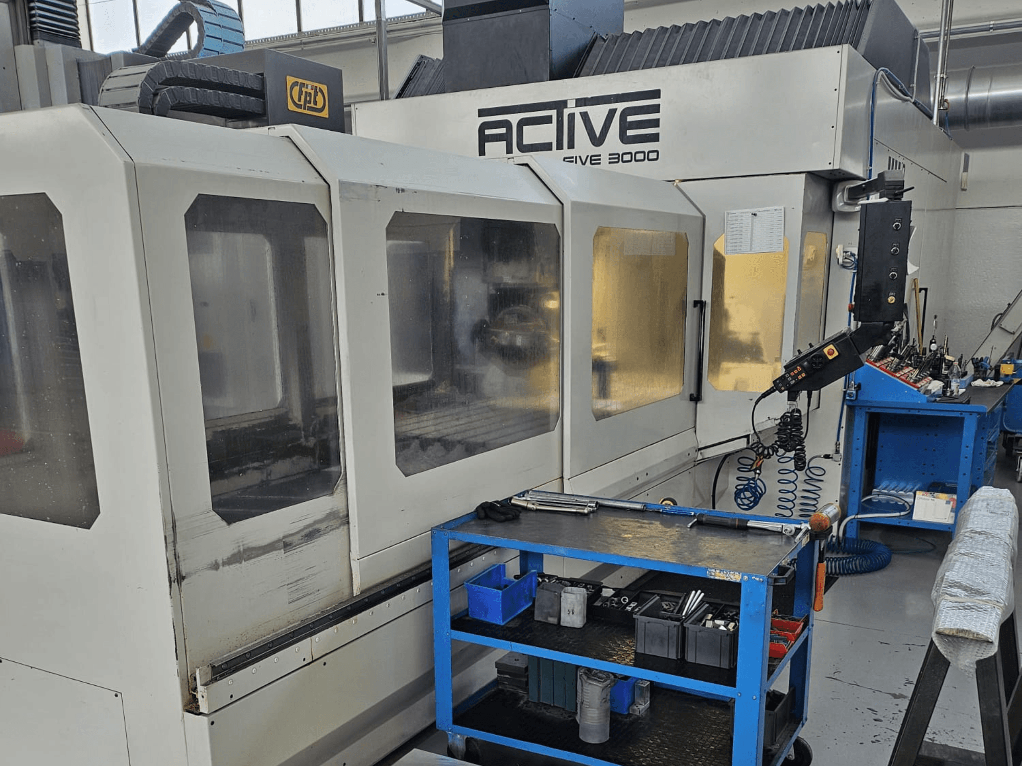 Aktiv 3000 CNC-maskin i en verkstad, framifrån med verktyg på en blå vagn, synliga kontroller och arbetsområde inuti.