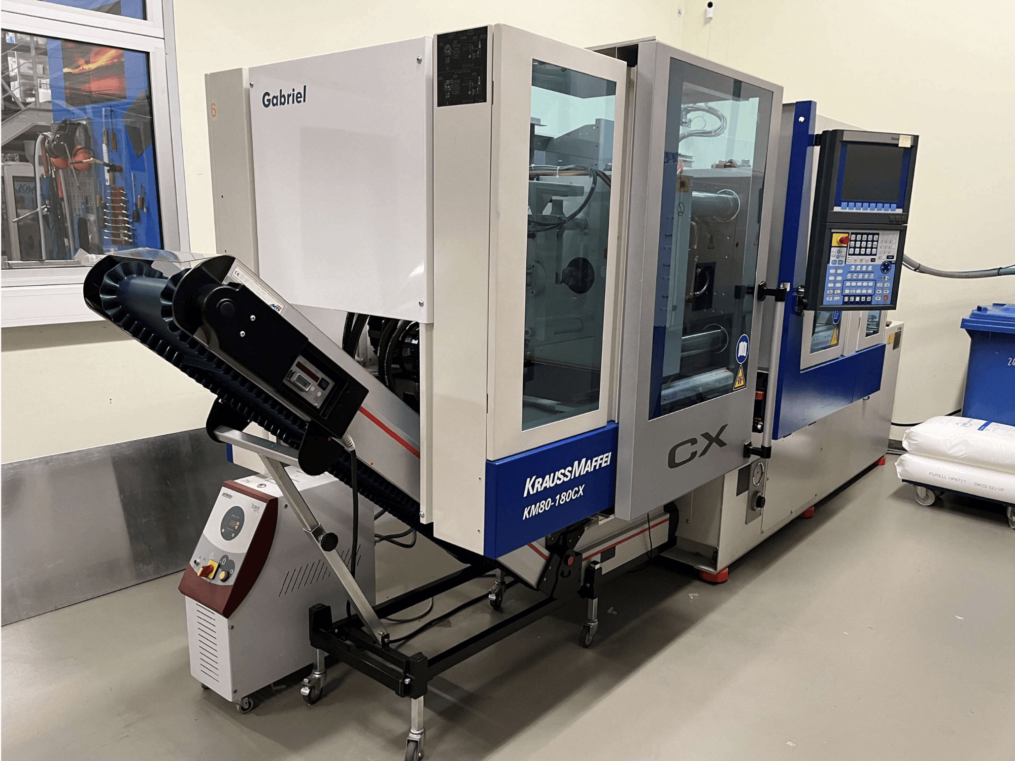 Hydraulisk formsprutningsmaskin Krauss Maffei KM 80-180 CX, framifrån, med kontrollpanel och transportsystem.