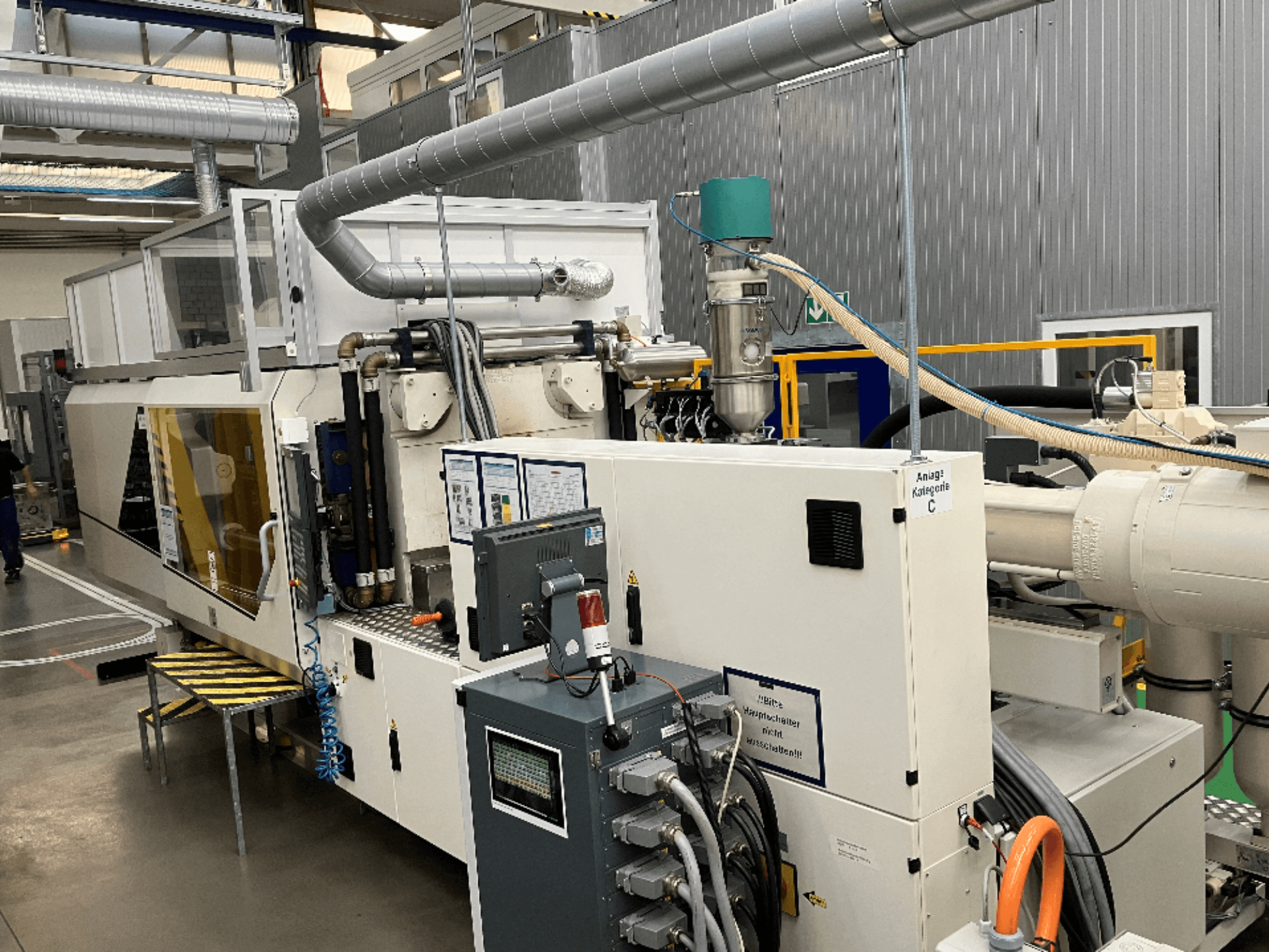 Formsprutningsmaskin, vy framifrån, med märkesetikett, kontrollpanel och synliga slangar i en fabriksmiljö.