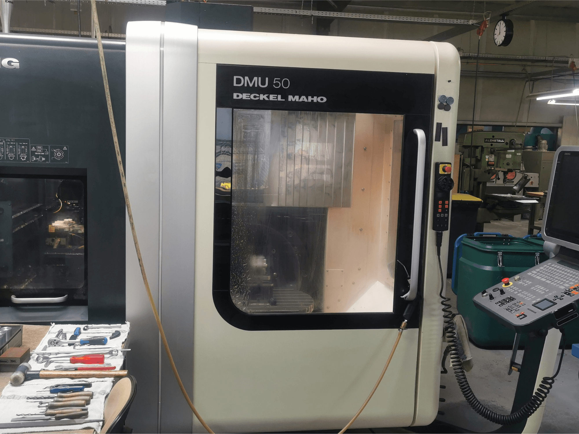 DMU 50 deckel maho CNC-maskin sedd framifrån, med kontrollpanel och transparent dörr. Verktyg finns i närheten.
