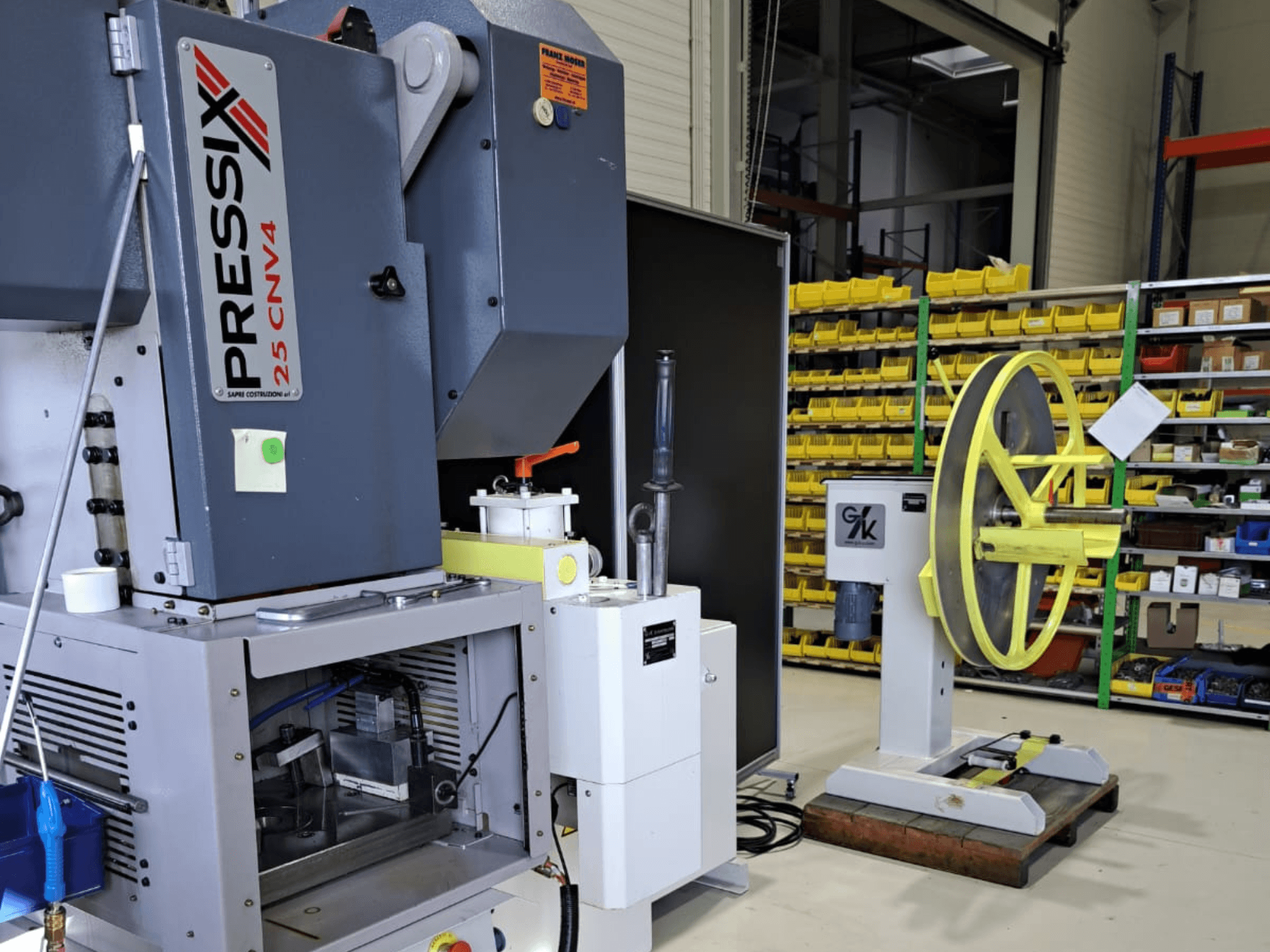 Industriell maskinuppställning med PRESSIX 25 CNV4 till vänster och ett gult hjul monterat på en grå bas till höger, framifrån.