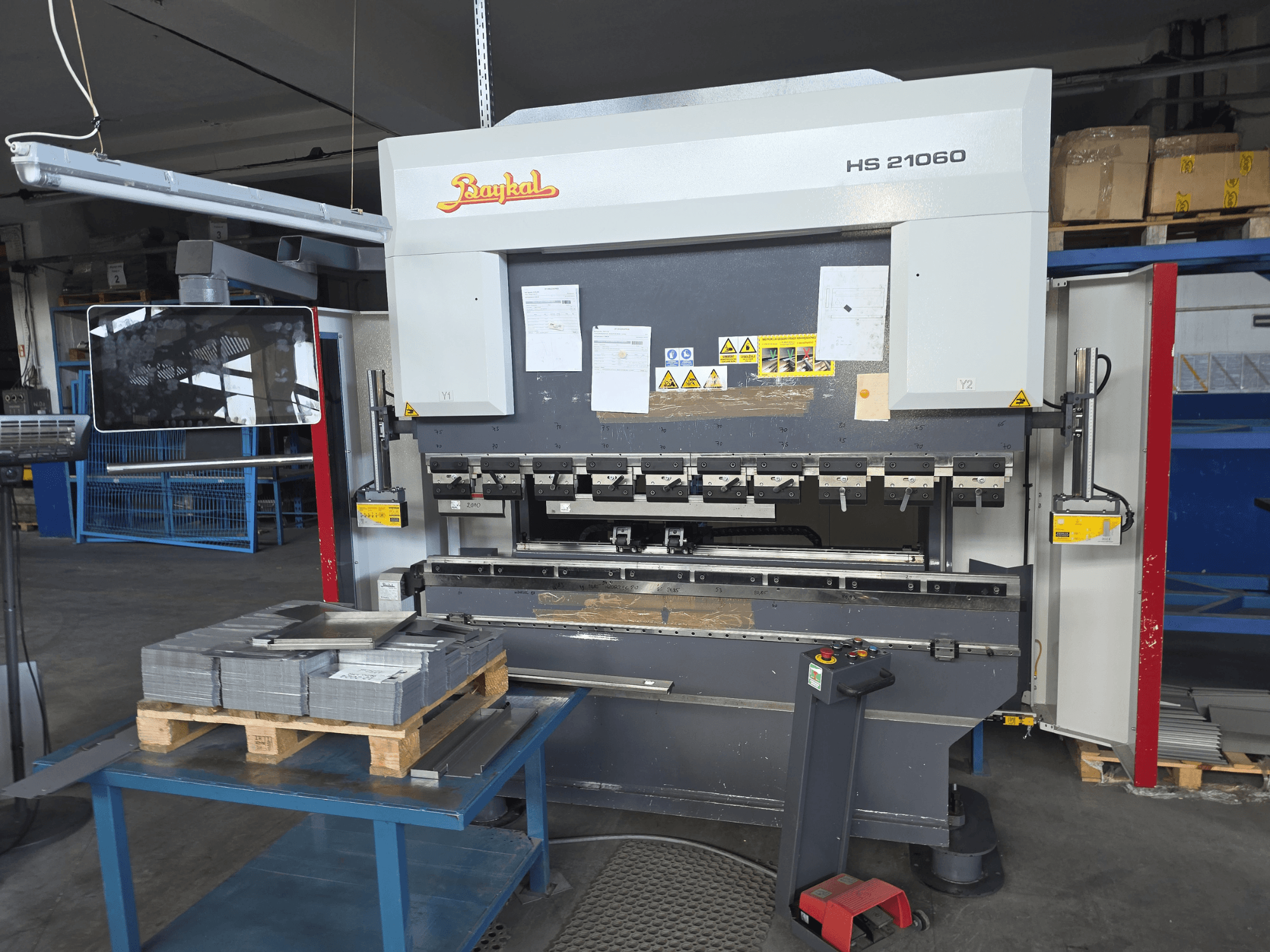 En Baykal APHS 21060 kantpress i en verkstad, sidovy som visar kontrollpanelen, staplade metallplåtar och säkerhetsskyltar.